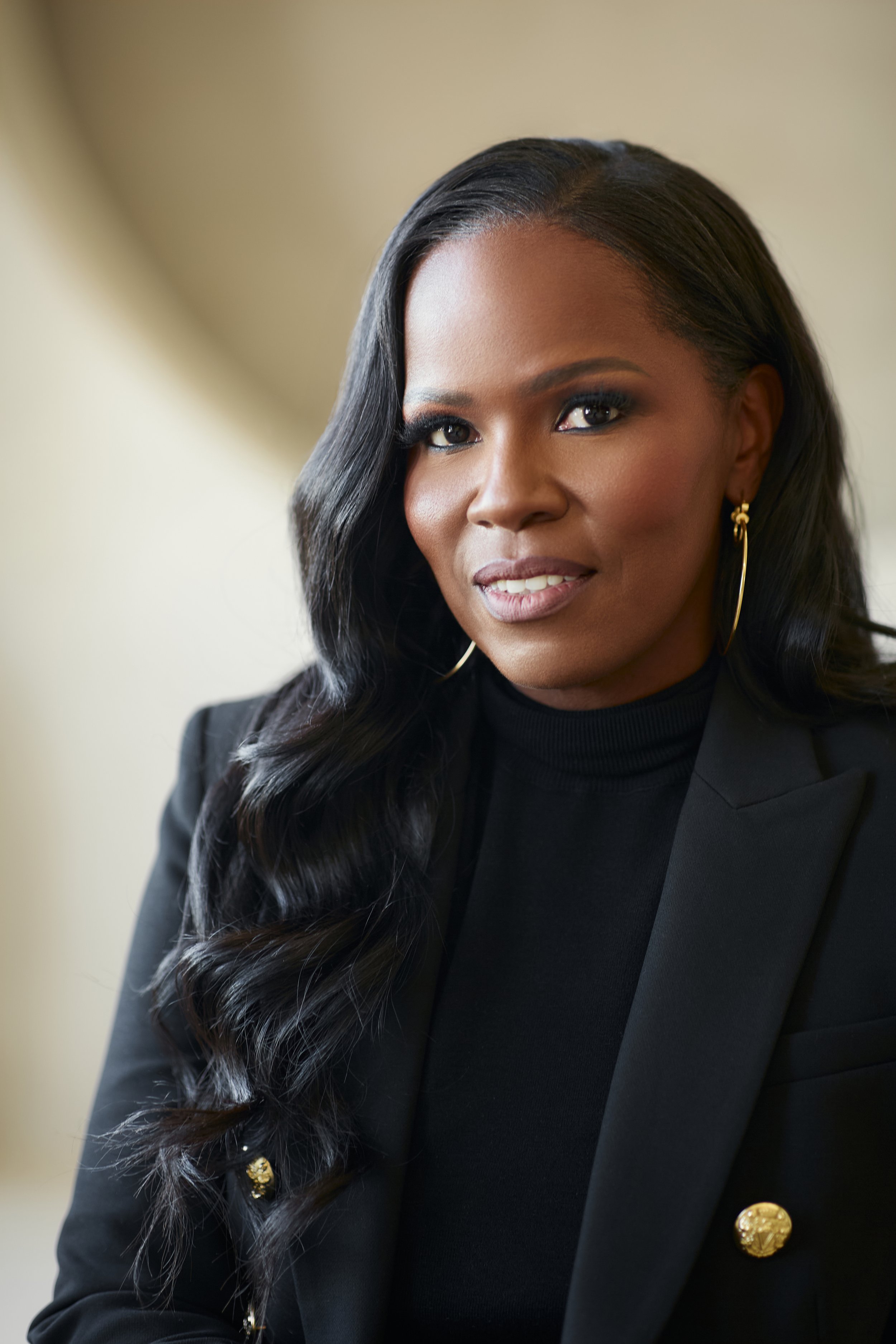 A woman with long, wavy black hair wearing gold hoop earrings and a black blazer with gold buttons, smiling softly at the camera.