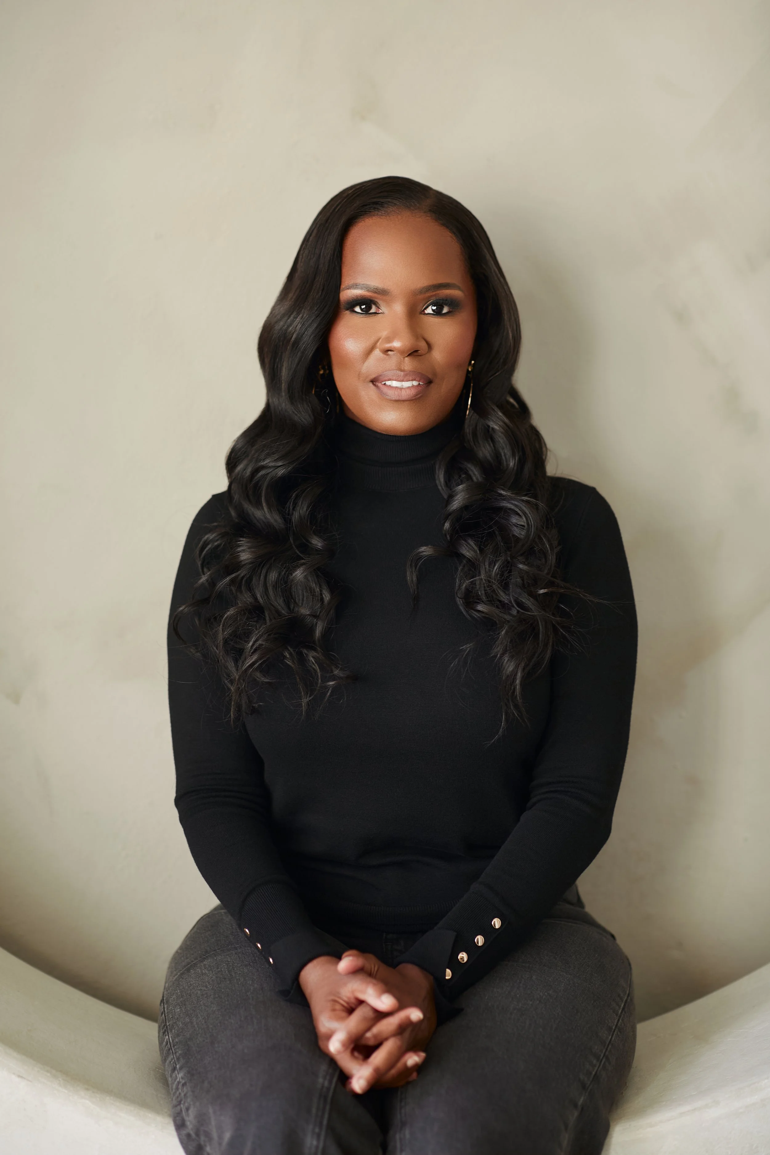 A woman with long, wavy black hair sitting in front of a light-colored wall, wearing a black turtleneck sweater and gray pants.