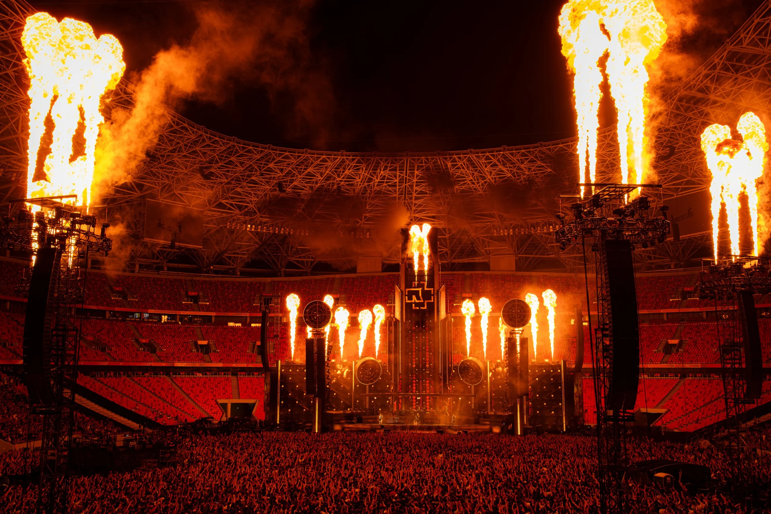A stadium at night with pyrotechnics and flames shooting into the sky from the stage and surrounding structures.