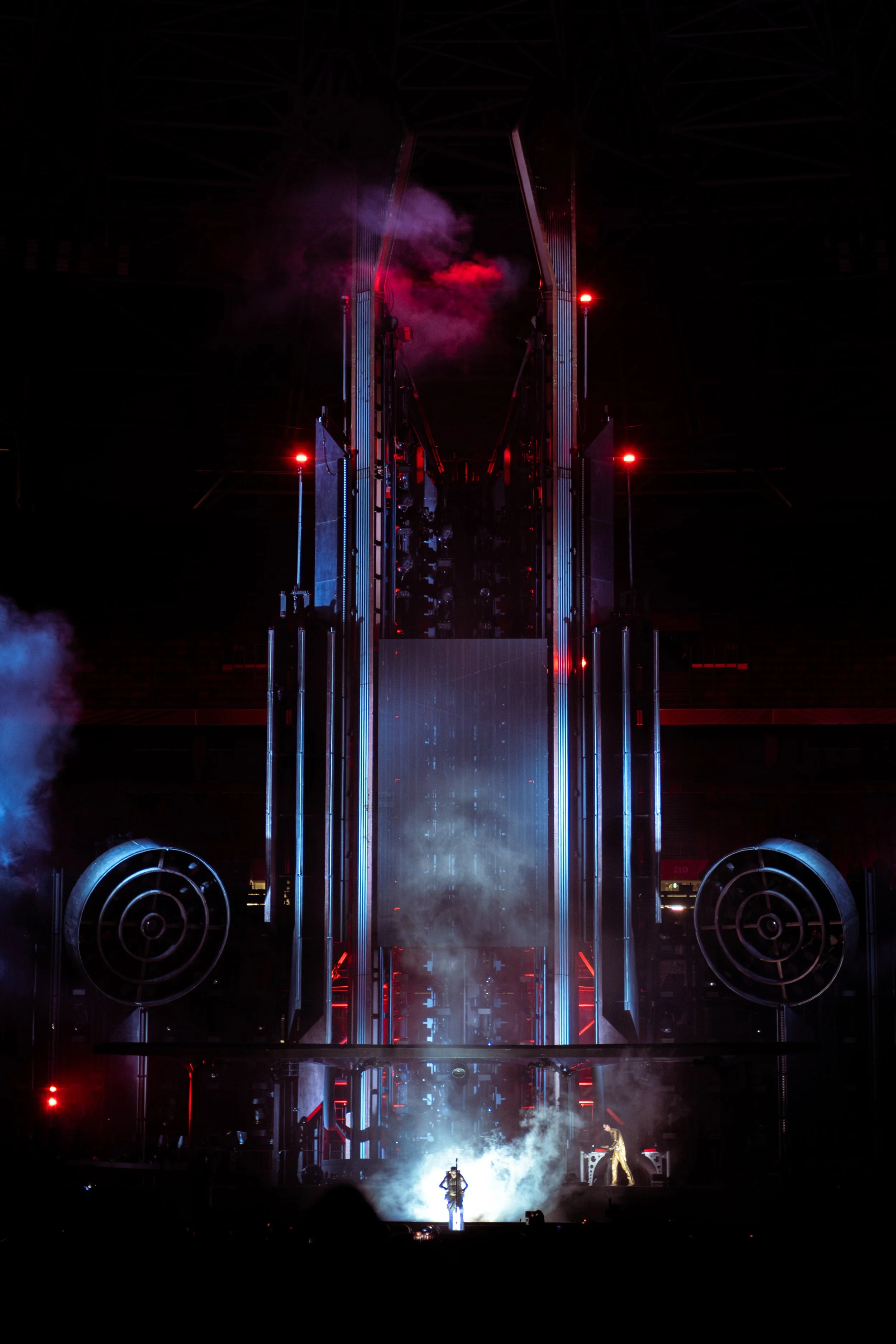Stage with performers, dramatic lighting, red and blue lights, smoke, and large speakers.