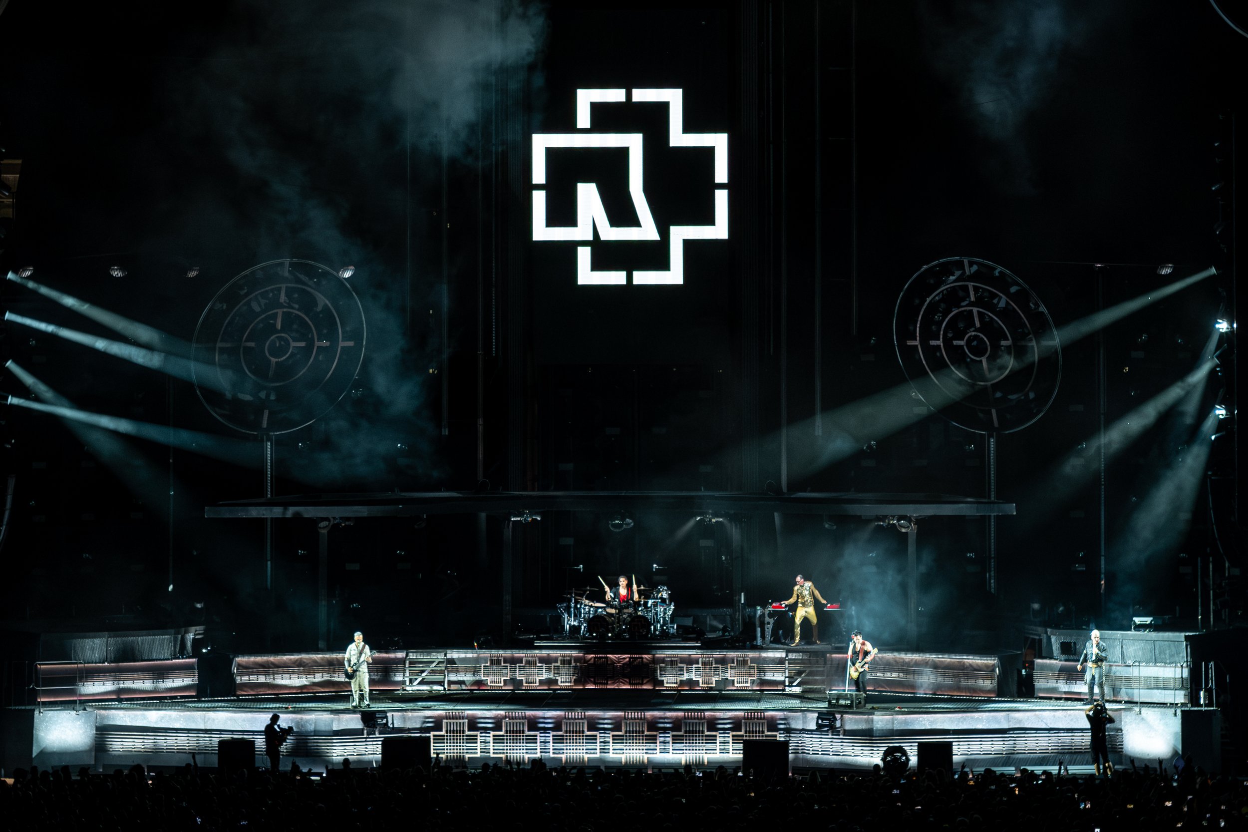 A live concert performance on a large stage with a band, musicians, and dancers. Bright lights and smoke effects create an energetic atmosphere. A large digital display with a logo is above the stage.