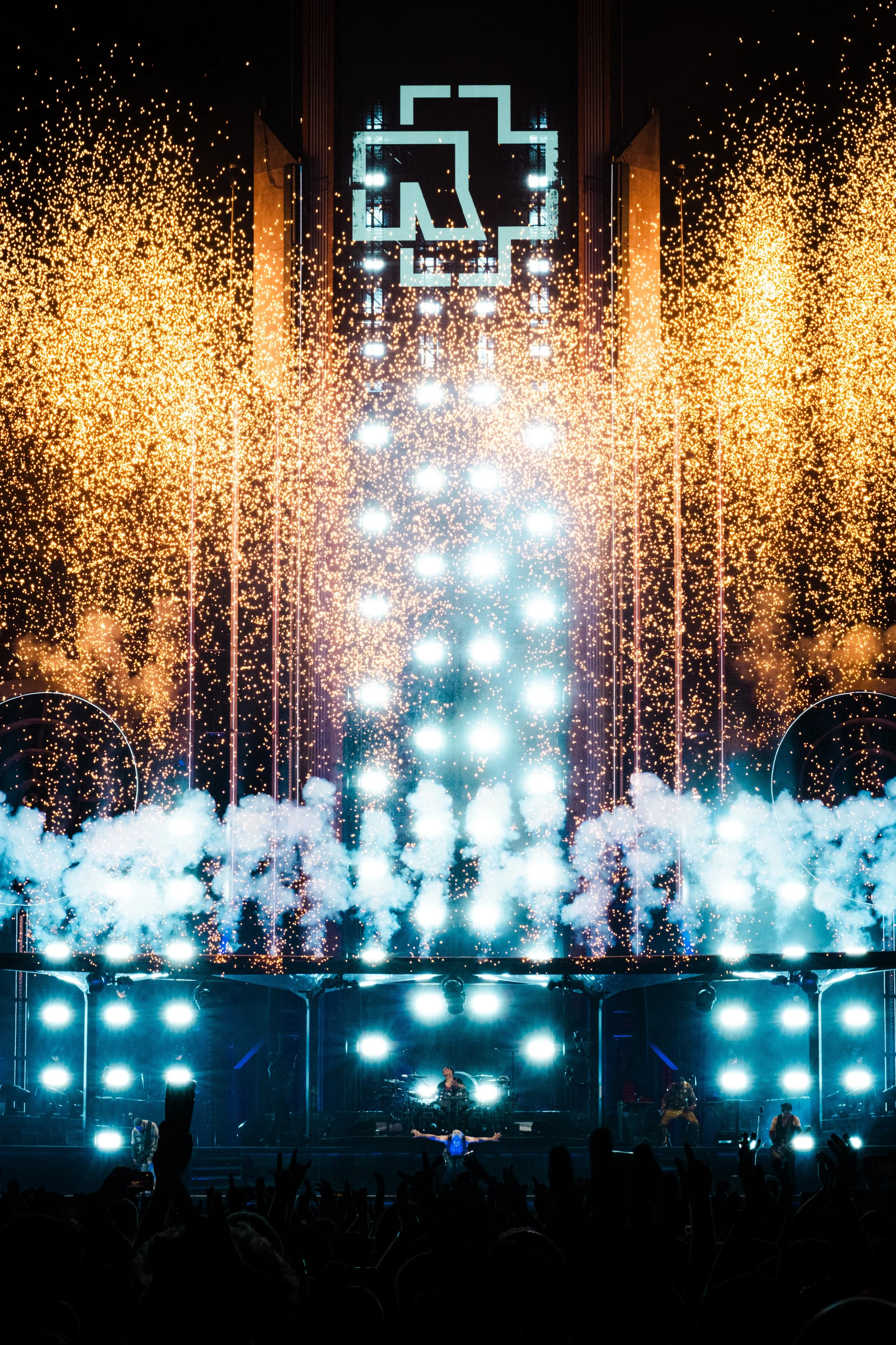 Concert stage with bright blue and white lighting, fireworks, and smoke, with silhouettes of audience members and performers.