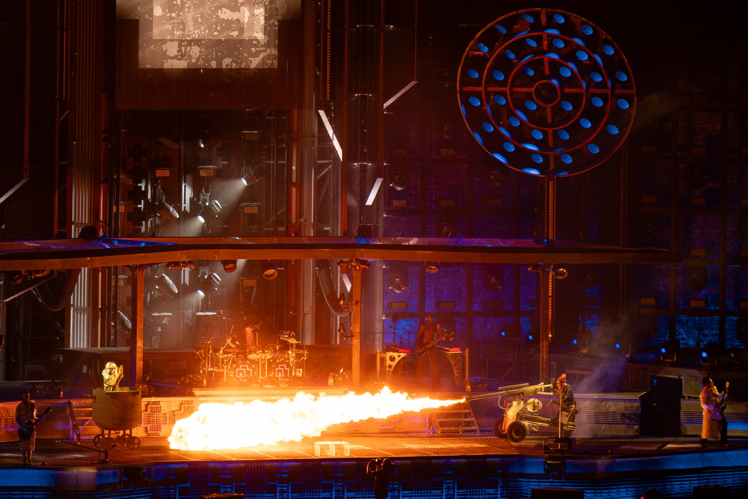 Stage scene with flames at the bottom, blue circular lighting structures, performers and crew, and screens showing abstract visuals, in a modern theater or concert setting.