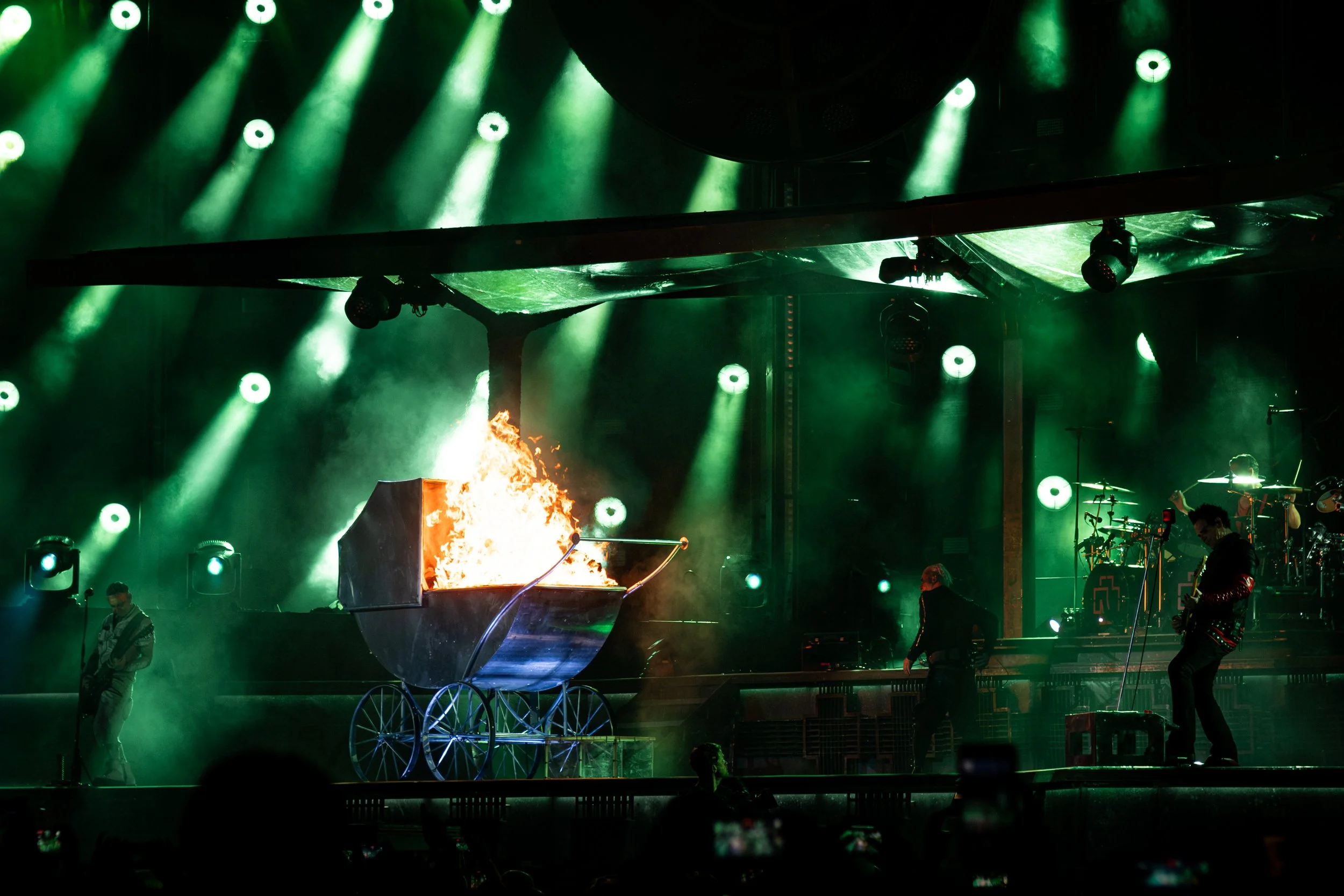 Stage performance with green lighting, a flaming prop, and performers dressed in costumes, resembling a circus or theatrical show.