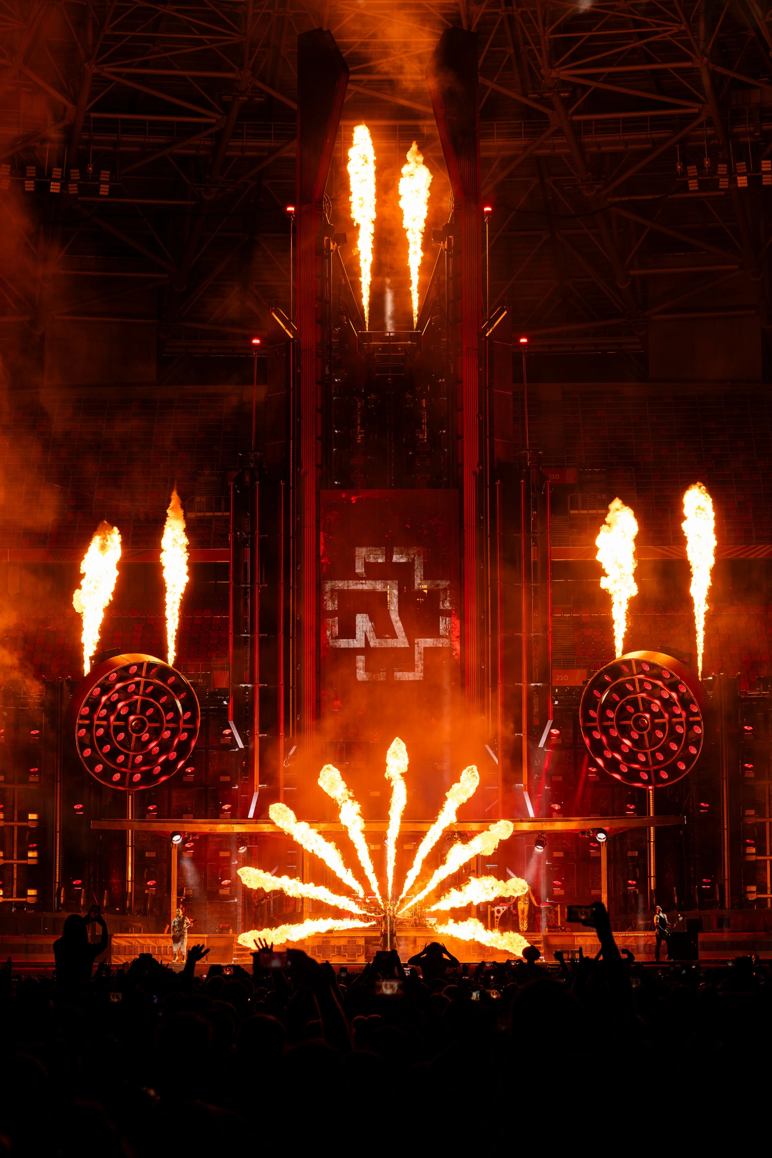 A concert scene at night with fireworks, flames, and a stage with structural metal framework. Audience silhouettes are visible in the foreground.