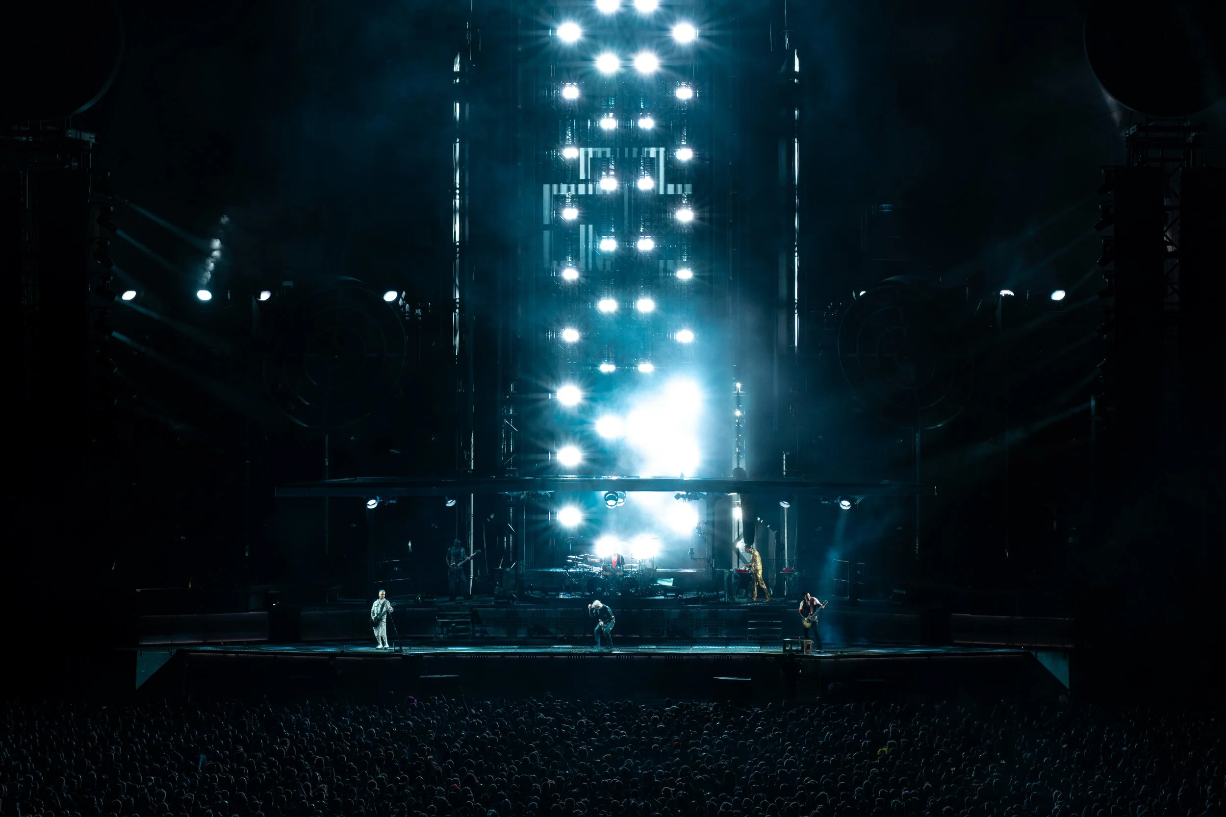 A large concert stage illuminated with bright white and blue lights, with a band performing and an audience watching in the dark.
