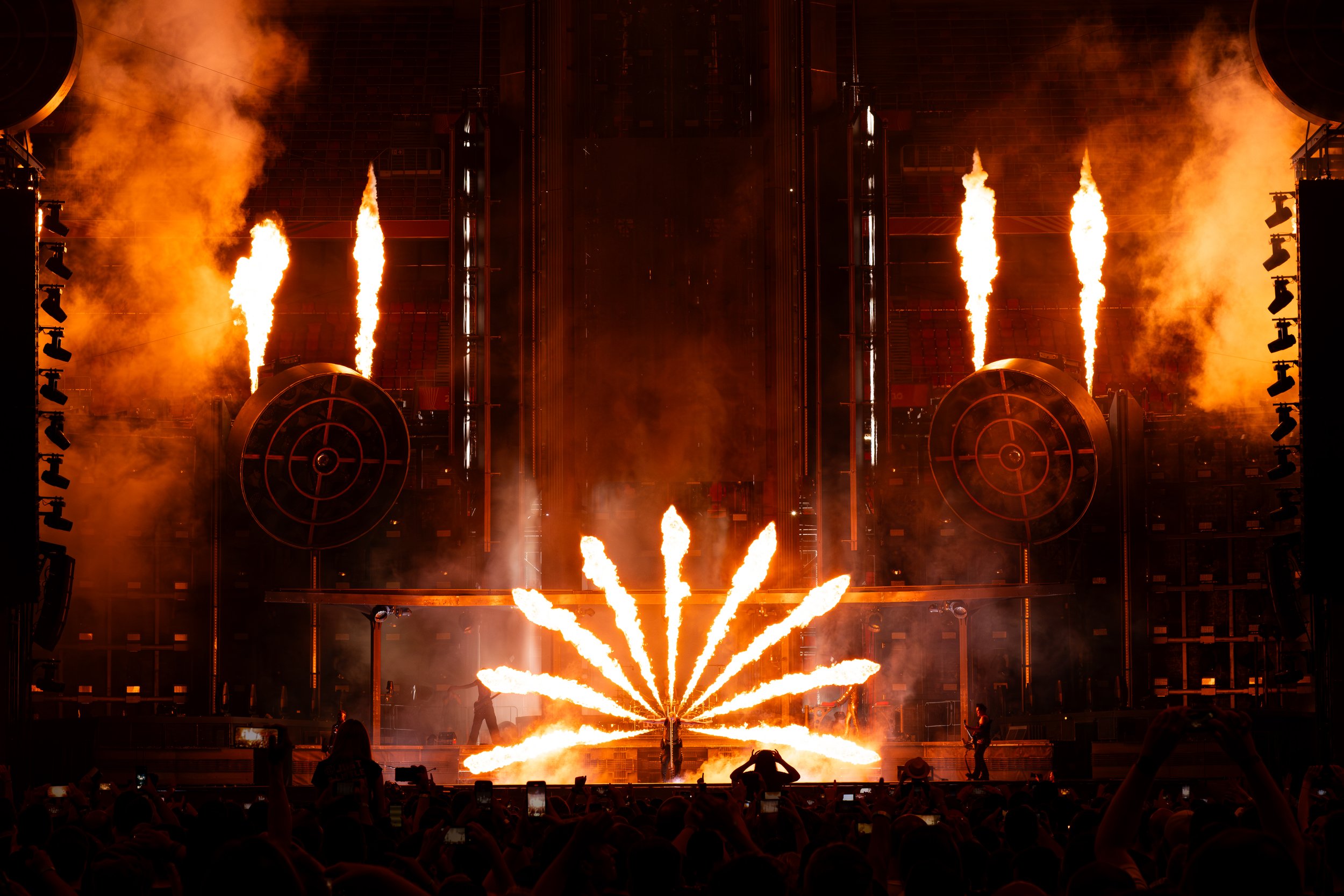 A concert stage with fire effects, including flames shooting up from the ground and large fire jets on the stage. Audience members are visible in the foreground watching the performance.