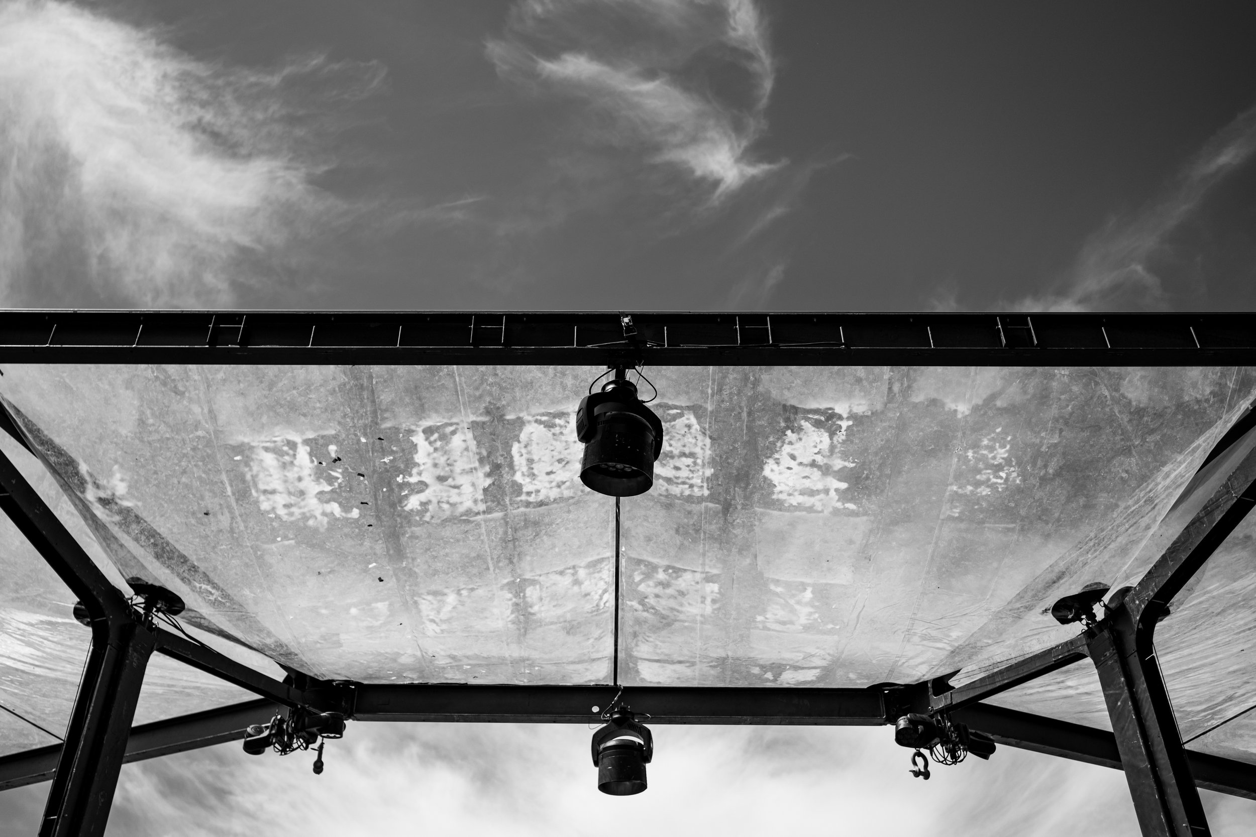 Black and white photo of stage lighting equipment mounted beneath a structure with a cloudy sky in the background.