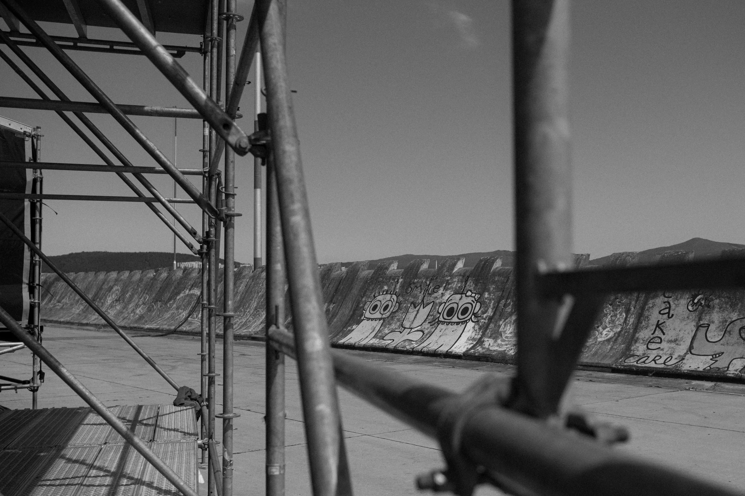 Black and white photo of a construction or maintenance site featuring metal scaffolding. In the background, graffiti art includes cartoon characters and text visible on a curved wall or surface, with clear mountains or hills in the distant landscape.