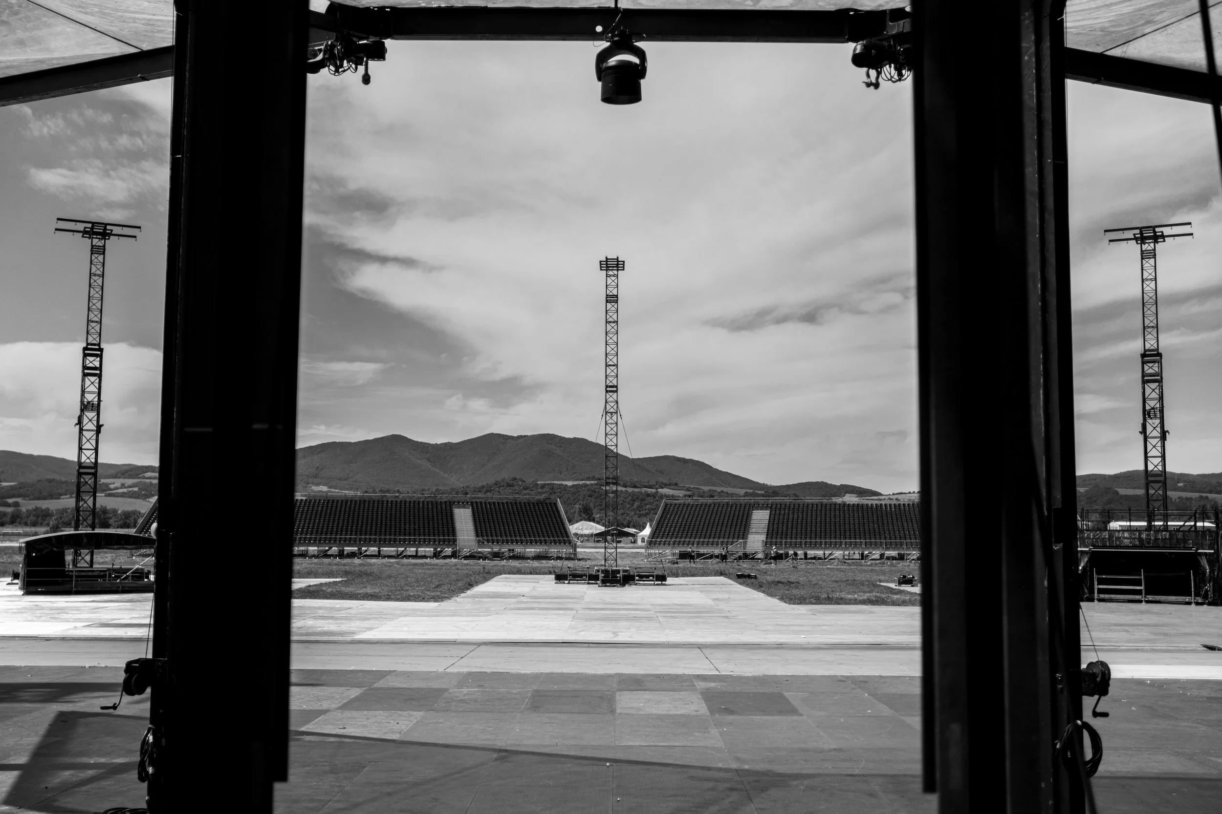 View from inside a large open-air stadium or sports arena, looking outward at three tall broadcast towers with mountains in the distance, black and white photography.