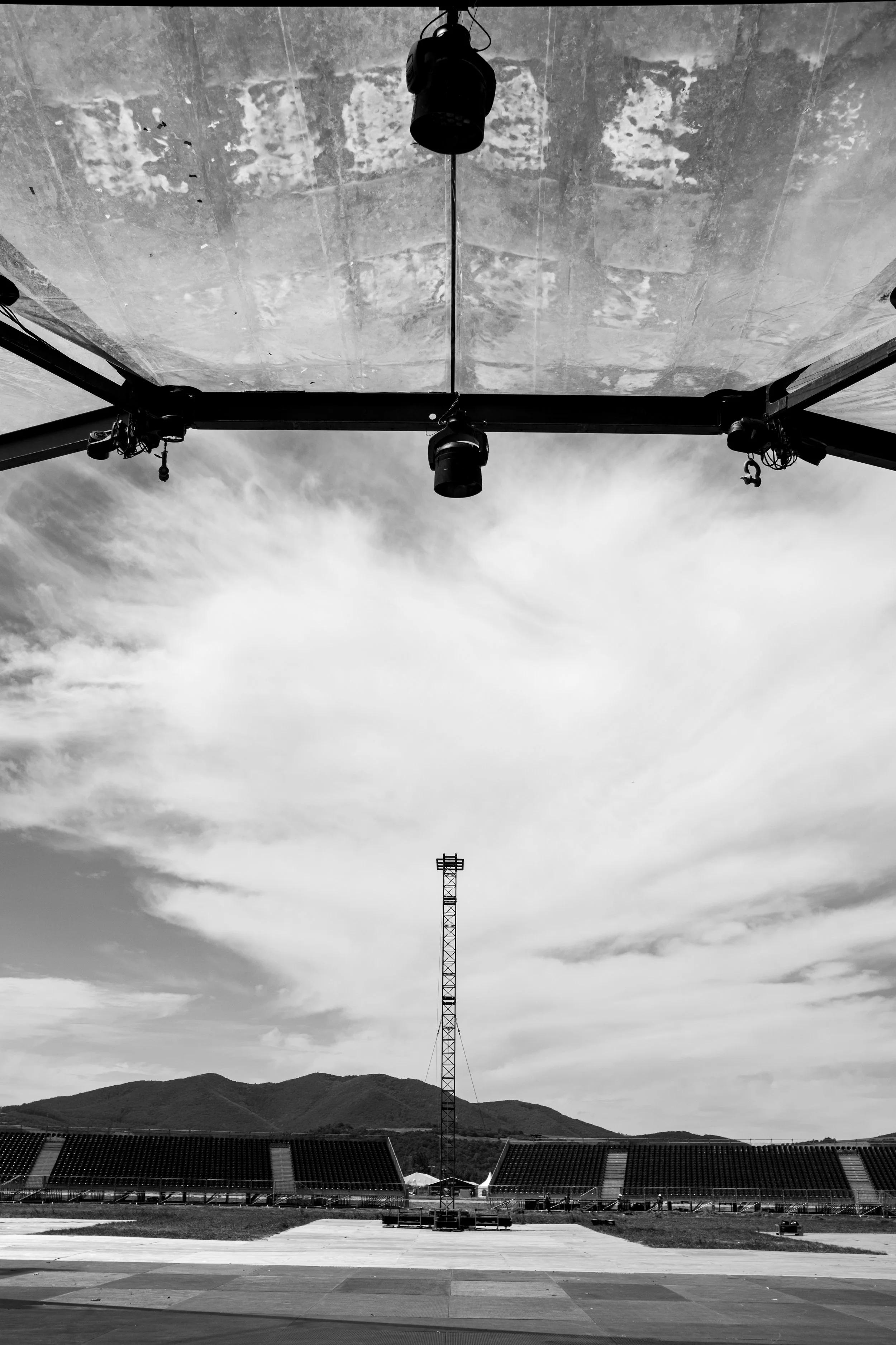 A black and white photo of a sports stadium with empty seats, a tall metal tower in the center, and a large overhead structure with mounted lights and equipment. Mountains are visible in the background under a cloudy sky.
