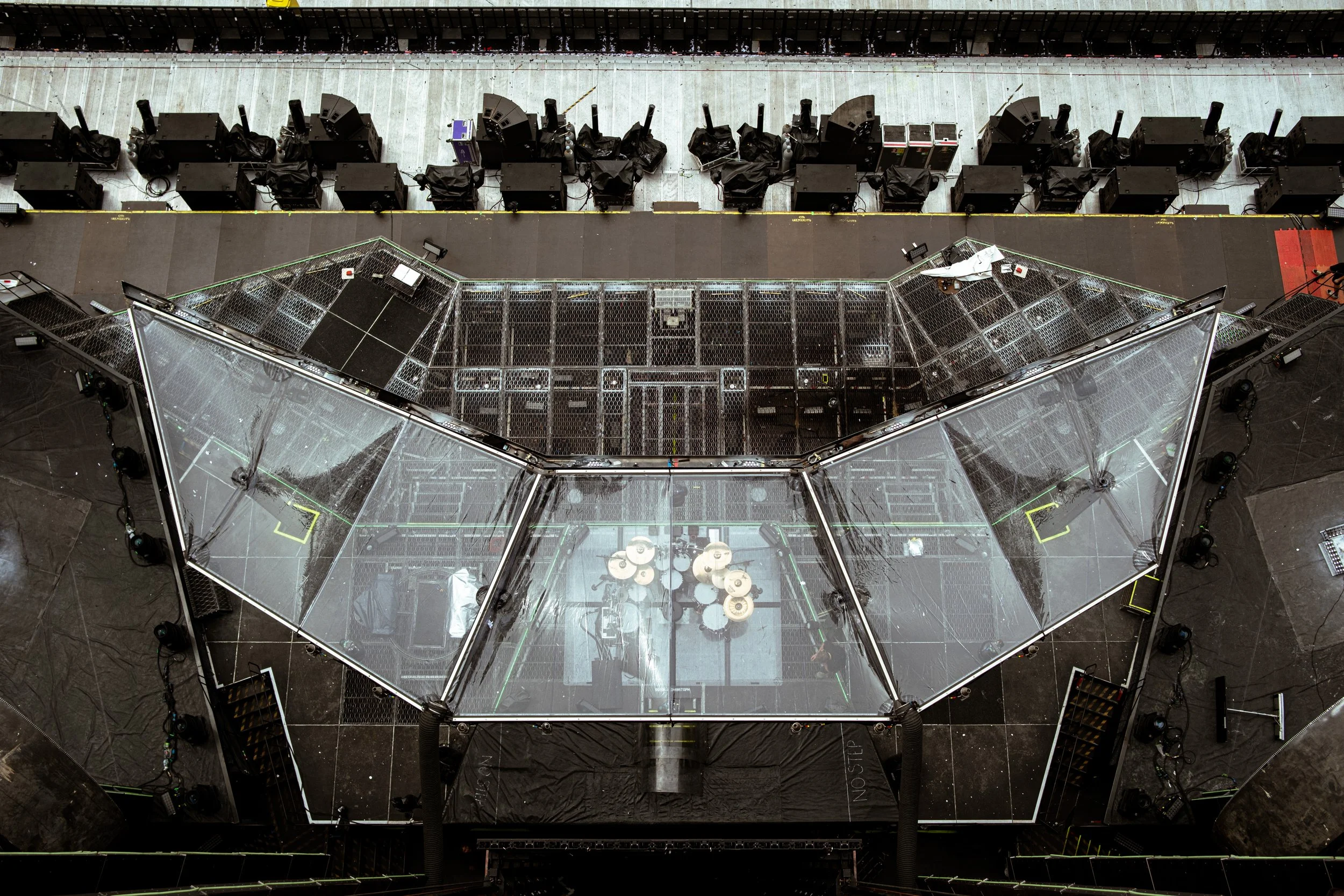Aerial view of a stage setup with clear panels, black equipment, and wine barrels inside the stage area, with black sound and lighting equipment around it.