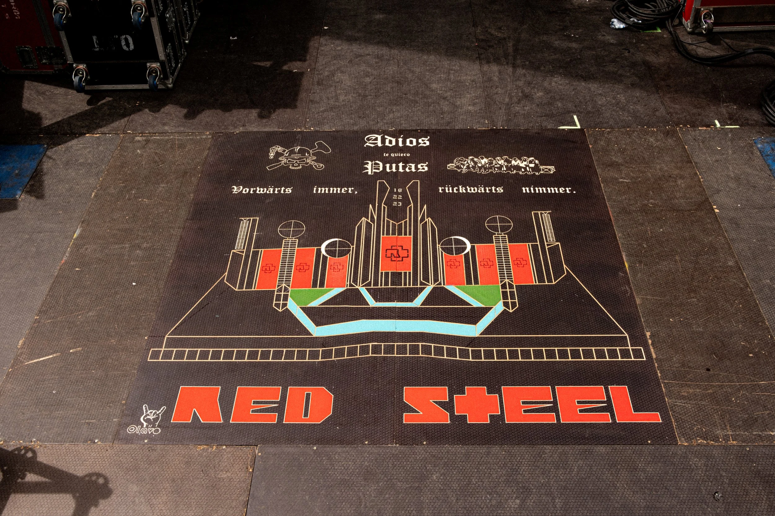 Floor mat with graffiti-style artwork and German text, including a depiction of a skull with a hat, in a setting with stacked equipment and cables.