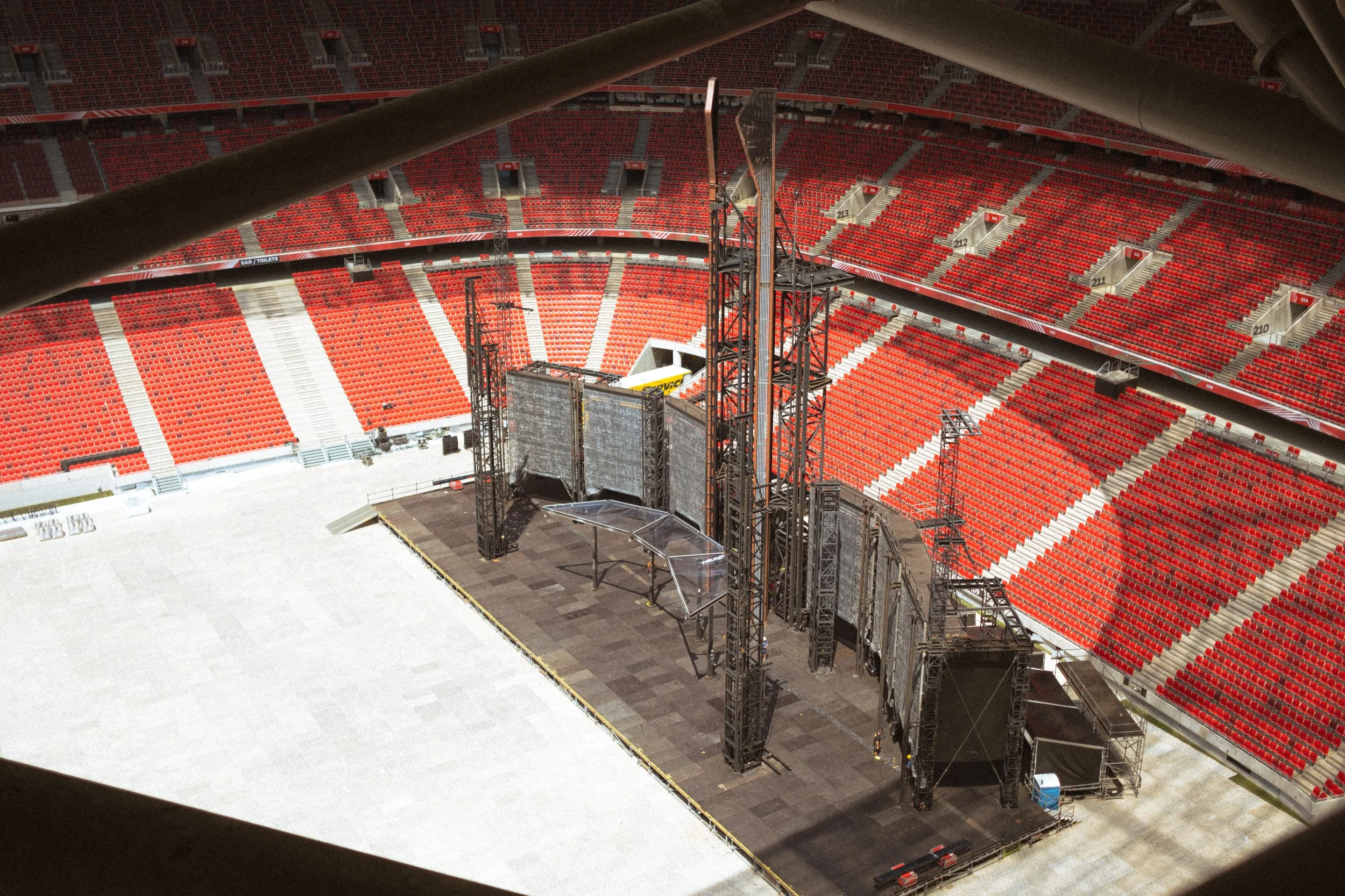 Interior of a large stadium with red seating, facing a stage with scaffolding and large screens under construction.