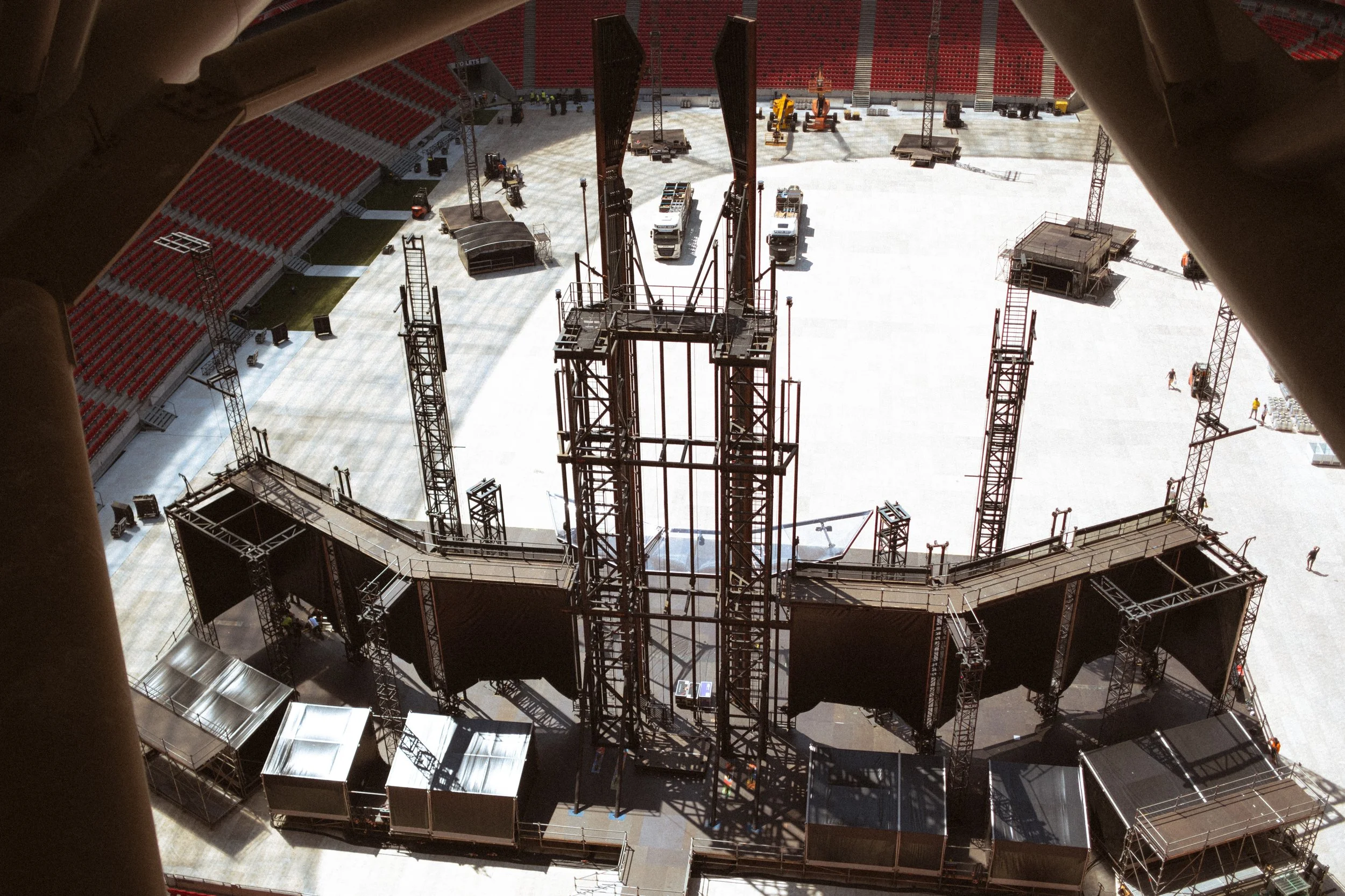 View from a high vantage point showing construction or setup inside an arena with large black speaker towers, metal scaffolding, trucks, and equipment, with empty red arena seats in the background.