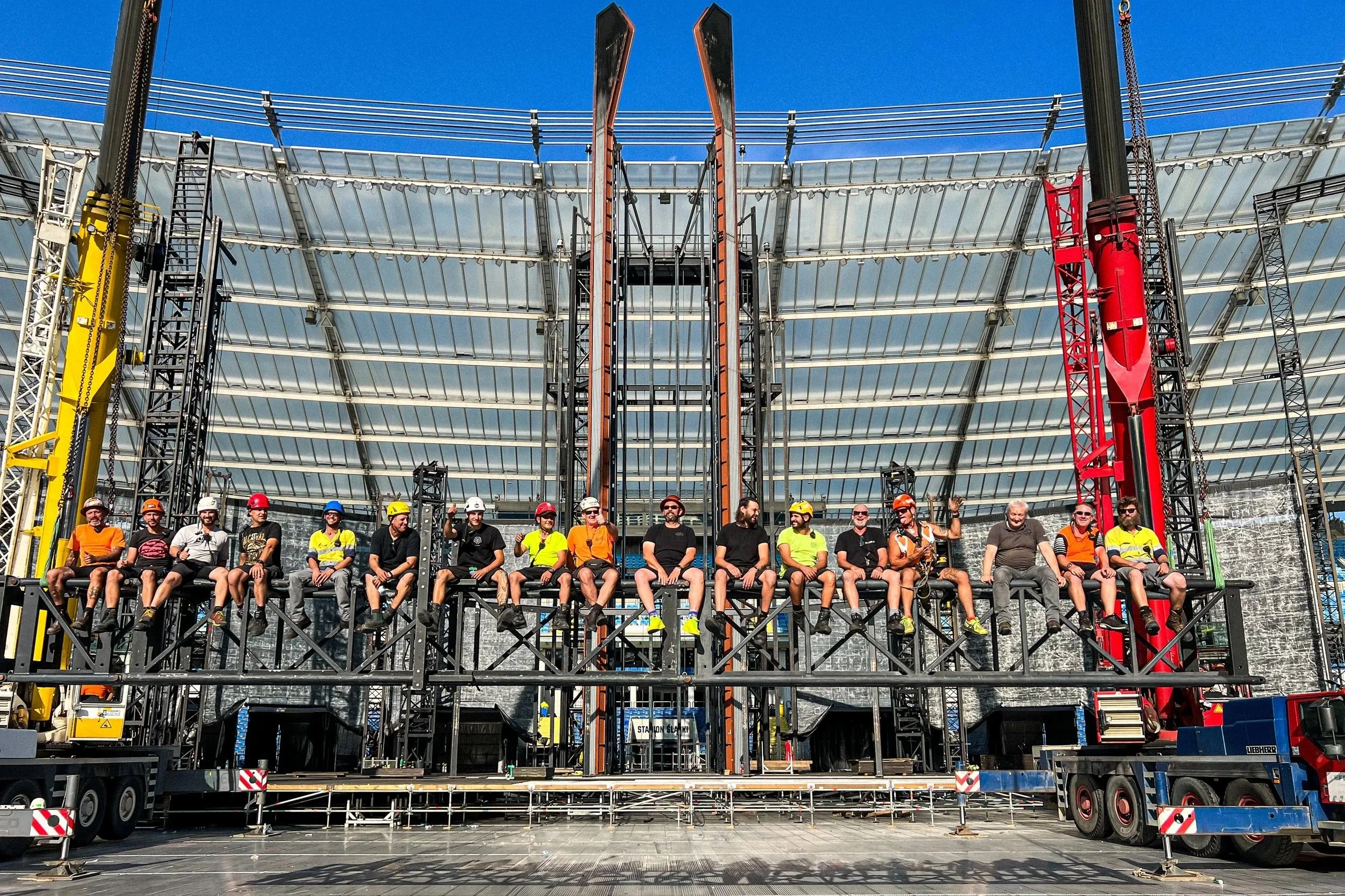 A group of people wearing safety gear, including helmets and harnesses, sitting on scaffolding in front of a large stage under construction. The stage has a metal framework and is being built with cranes and construction equipment.