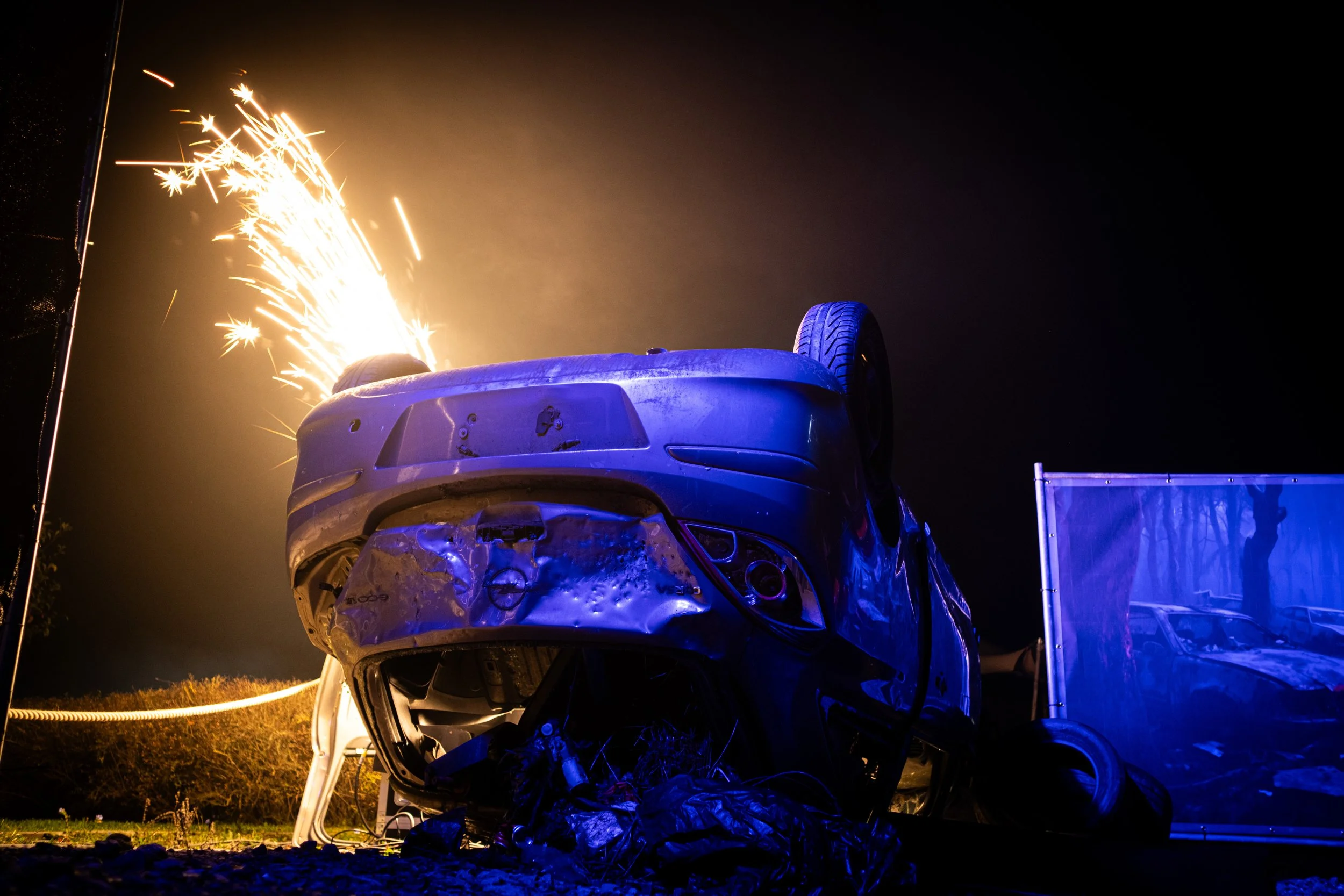 A car is overturned and on fire at night, with sparks and flames coming from the vehicle, which appears to be a damaged Opel with visible front-end destruction. The scene is illuminated by the fire, with a dark sky in the background.