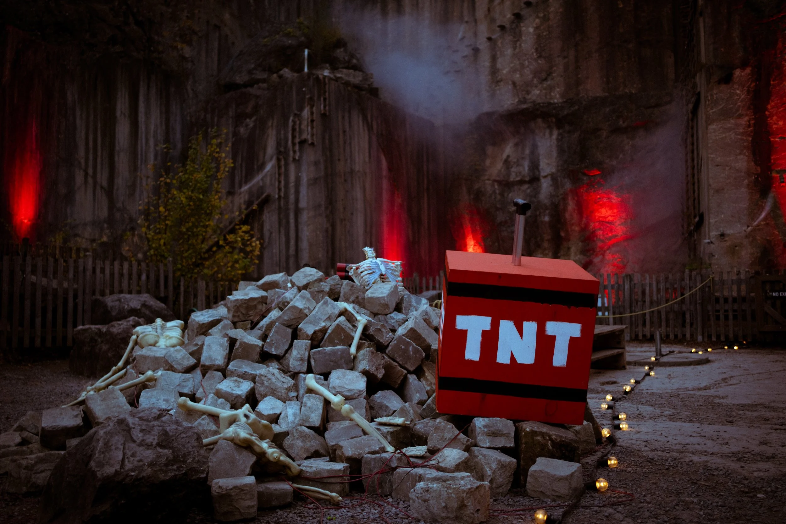 Creatively arranged pile of rocks and plastic bones in front of a steep rocky wall illuminated with red and white lights, with a red box labeled 'TNT' nearby, and a row of small lights along the ground.