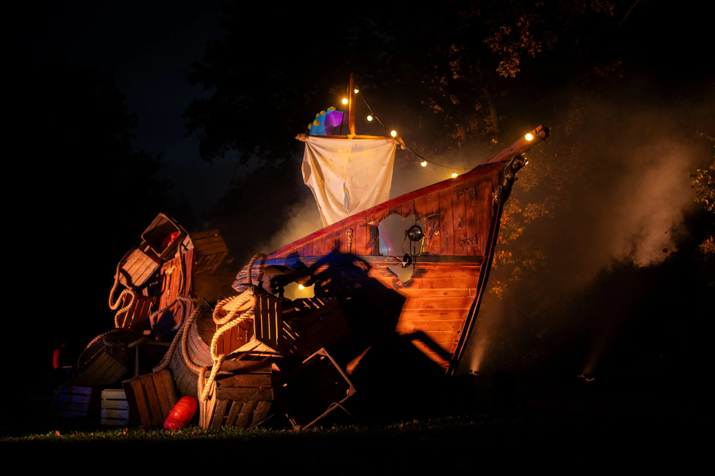 Nighttime scene featuring a wooden ship deck with pirate-themed decorations, illuminated by string lights, with trees in the background and mist or fog around the deck.