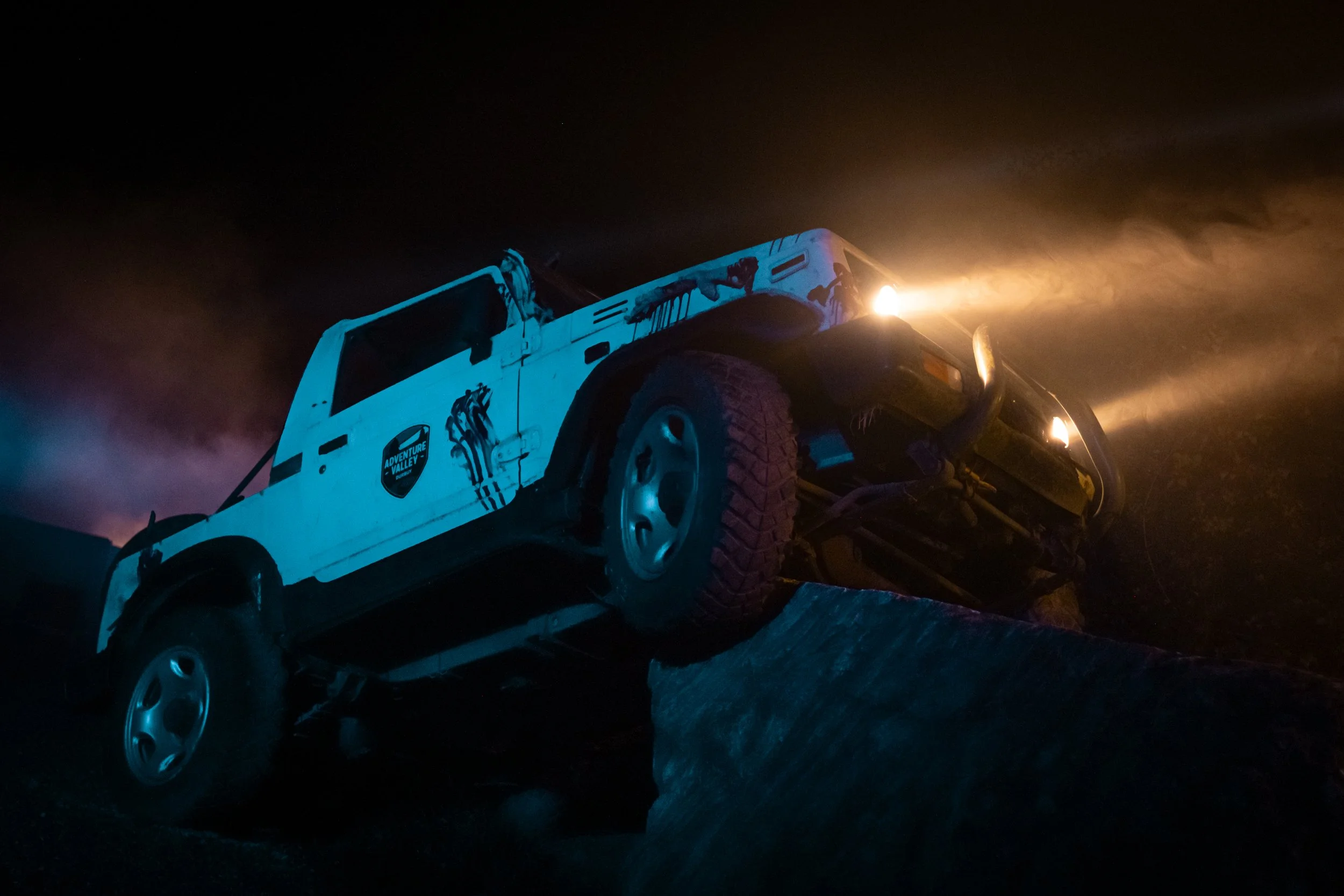A white off-road vehicle with decals, tilted at an angle on a rocky terrain at night, with bright light and dust clouds in the background.