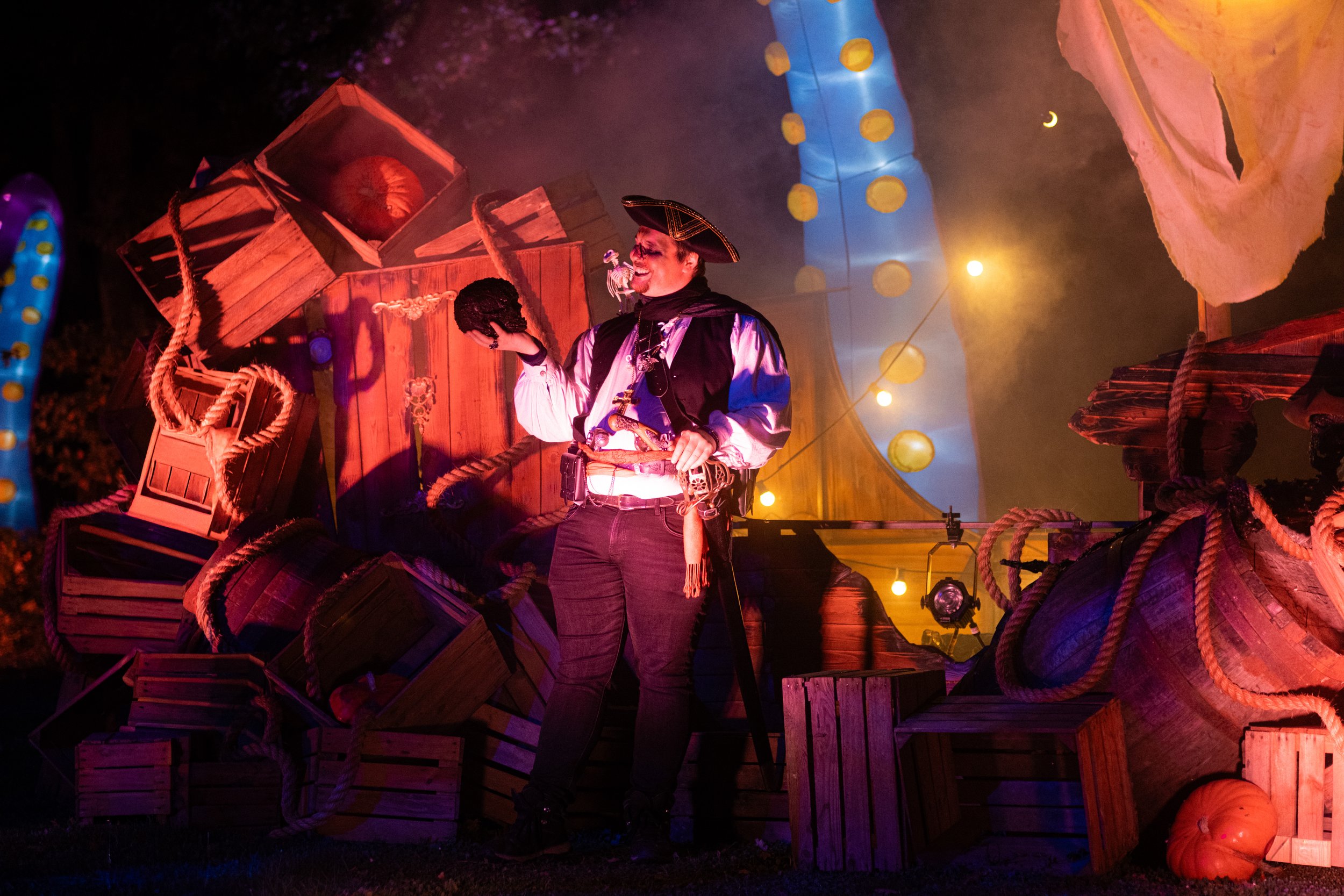 A person dressed as a pirate holding a skull prop, standing in a theatrical or Halloween setting with decorated structures, Halloween lights, and pumpkins.