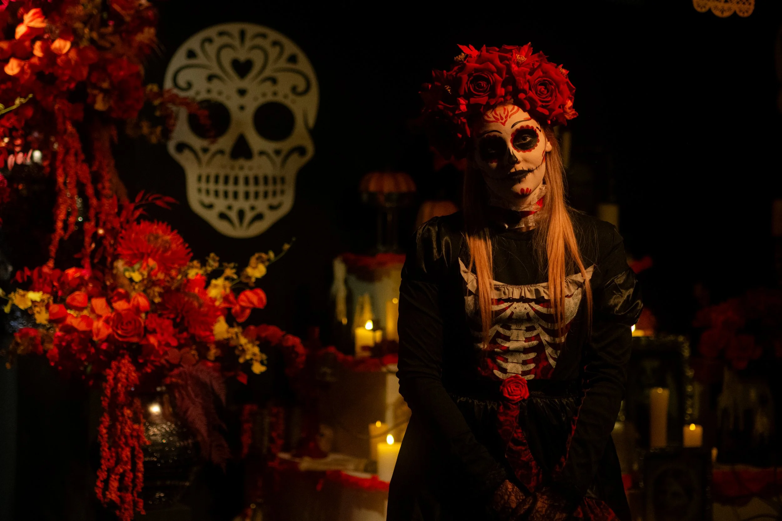 A person dressed in traditional Mexican Day of the Dead costume, painted face resembling a sugar skull, wearing a crown of red roses, standing in front of a decorated altar with candles, flowers, and sugar skulls.