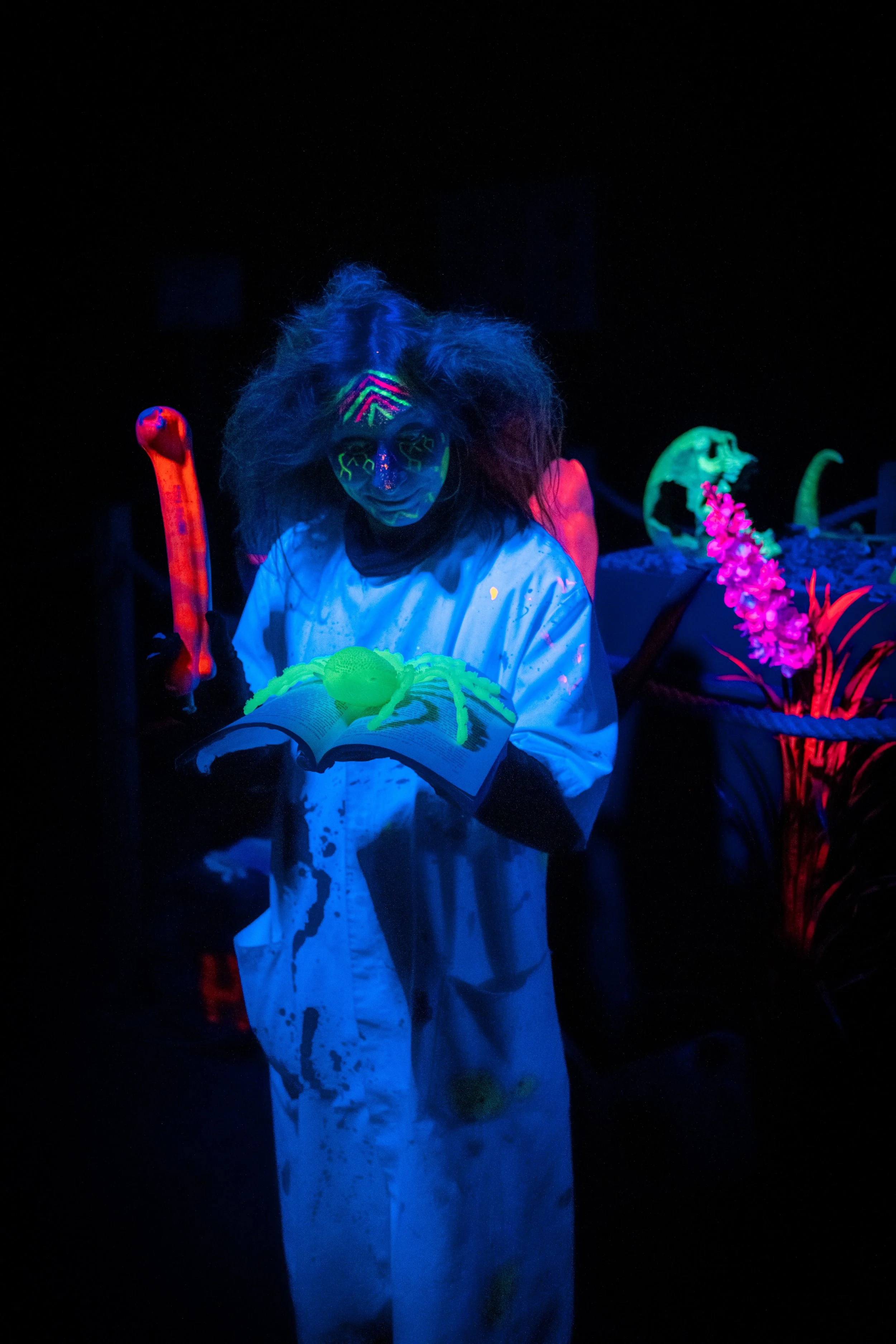 Person in glow-in-the-dark makeup and clothing holding a book, surrounded by glowing artificial flowers and dinosaur figures in a dark environment.