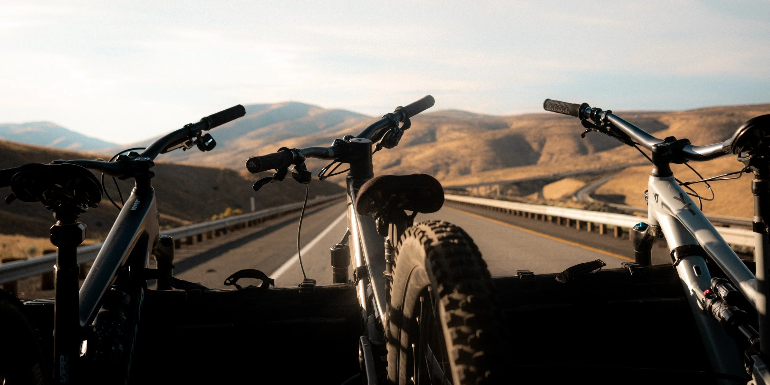 Two mountain bikes loaded in a truck bed, overlooking a winding highway through a hilly desert landscape during sunset in Oregon.