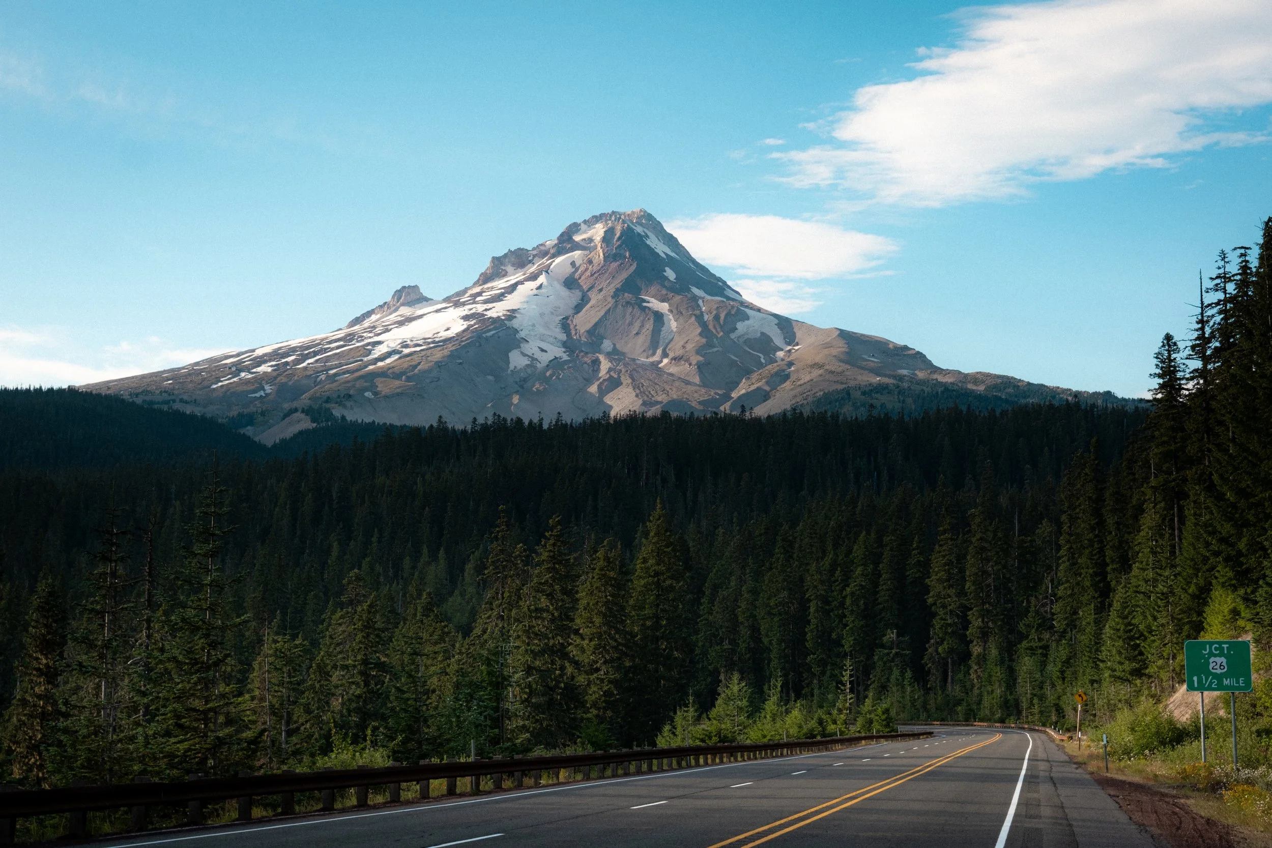 Scenic view of a mountain with snow patches, evergreen trees, and a winding road with a green highway sign indicating exit 26, 1/2 mile in Oregon.