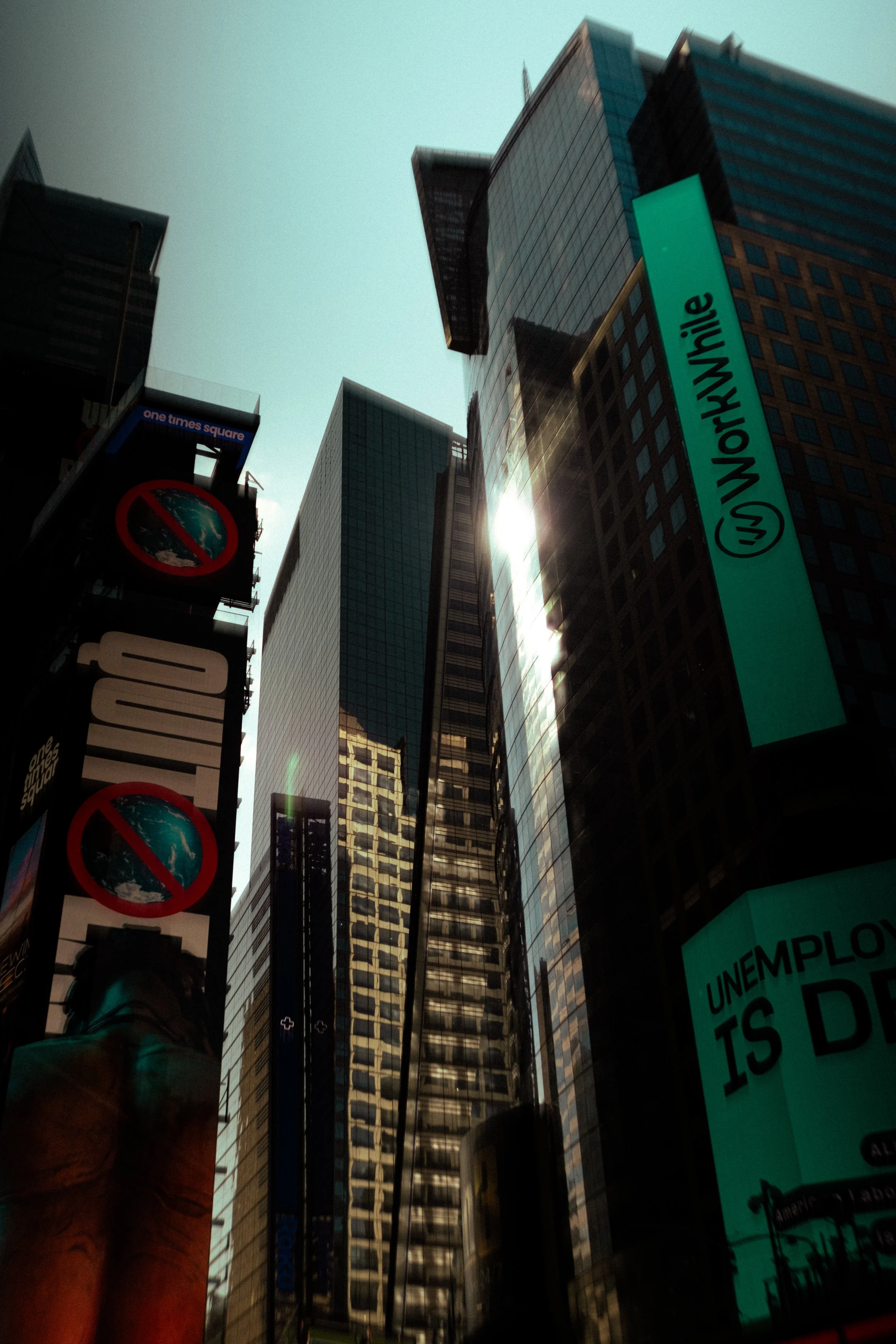 View of tall buildings and digital billboards in Times Square, New York City, with sunlight reflecting off glass facades.