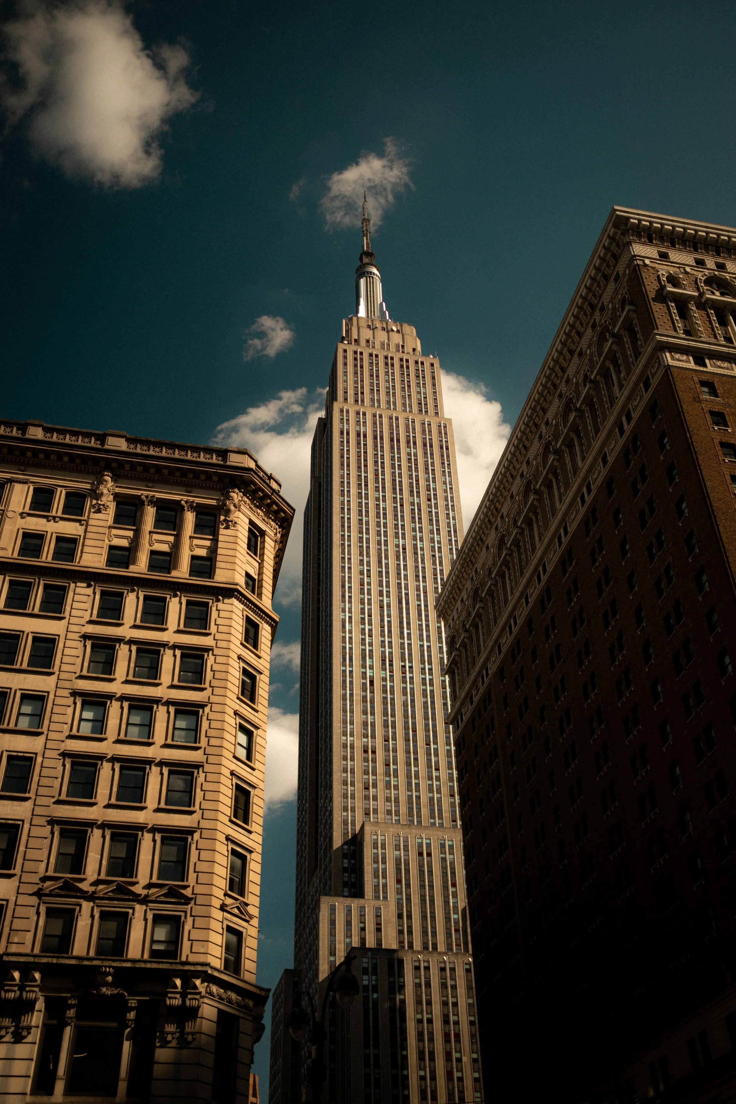 View of the Empire State Building in New York City, surrounded by other buildings against a blue sky with scattered clouds.