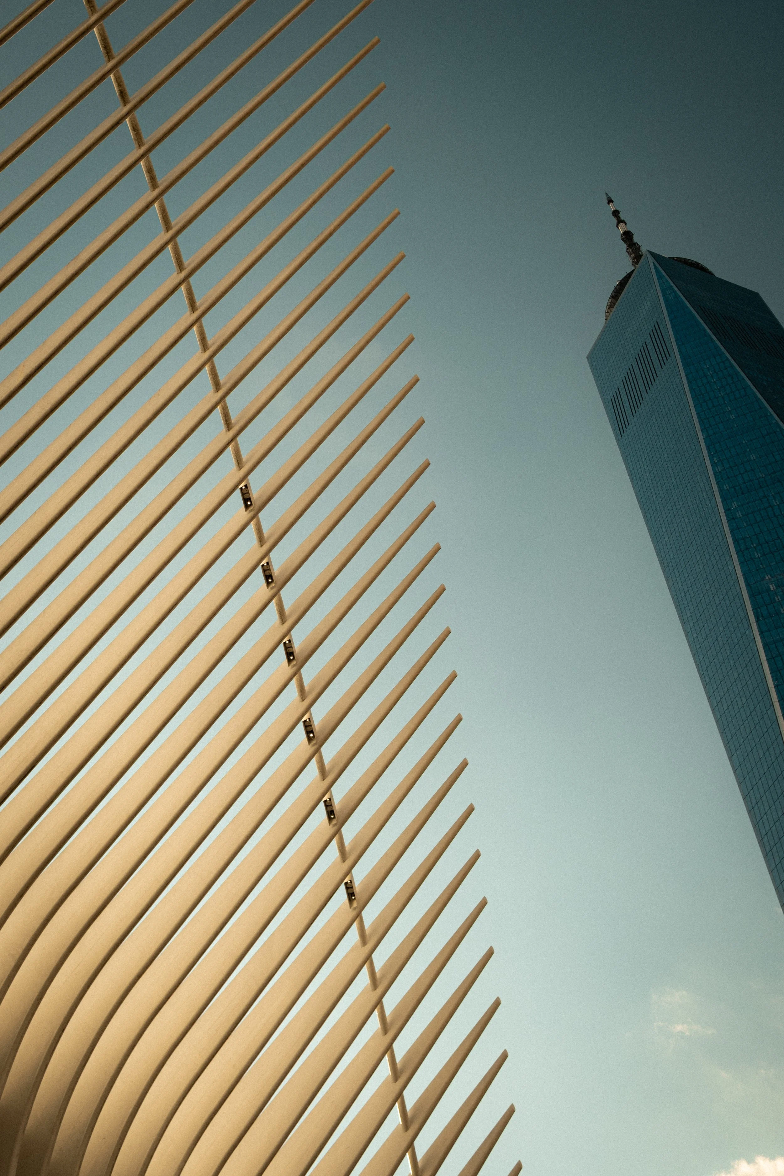 Looking up at a modern building with a sharp spire and reflective glass windows, with a curved metallic structure in the foreground against a clear sky in New York City.