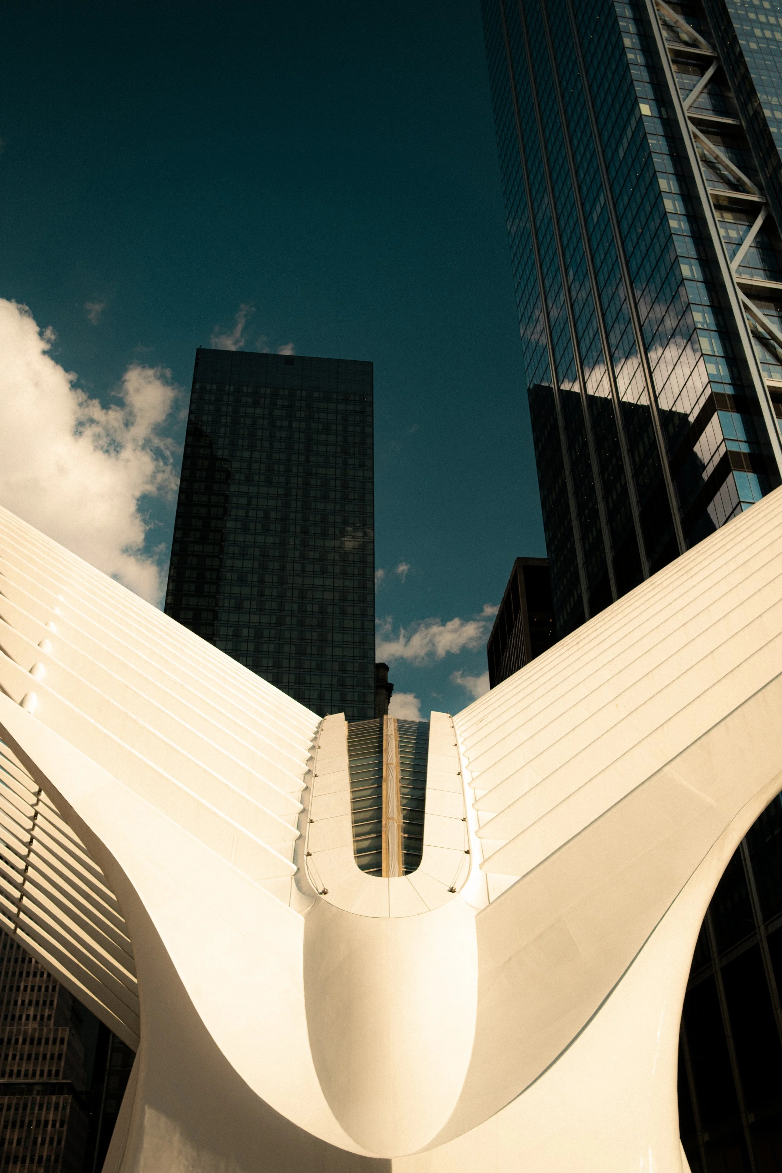 Modern architectural sculpture with a white curving form and a central opening, set against tall glass skyscrapers and a partly cloudy sky in New York City