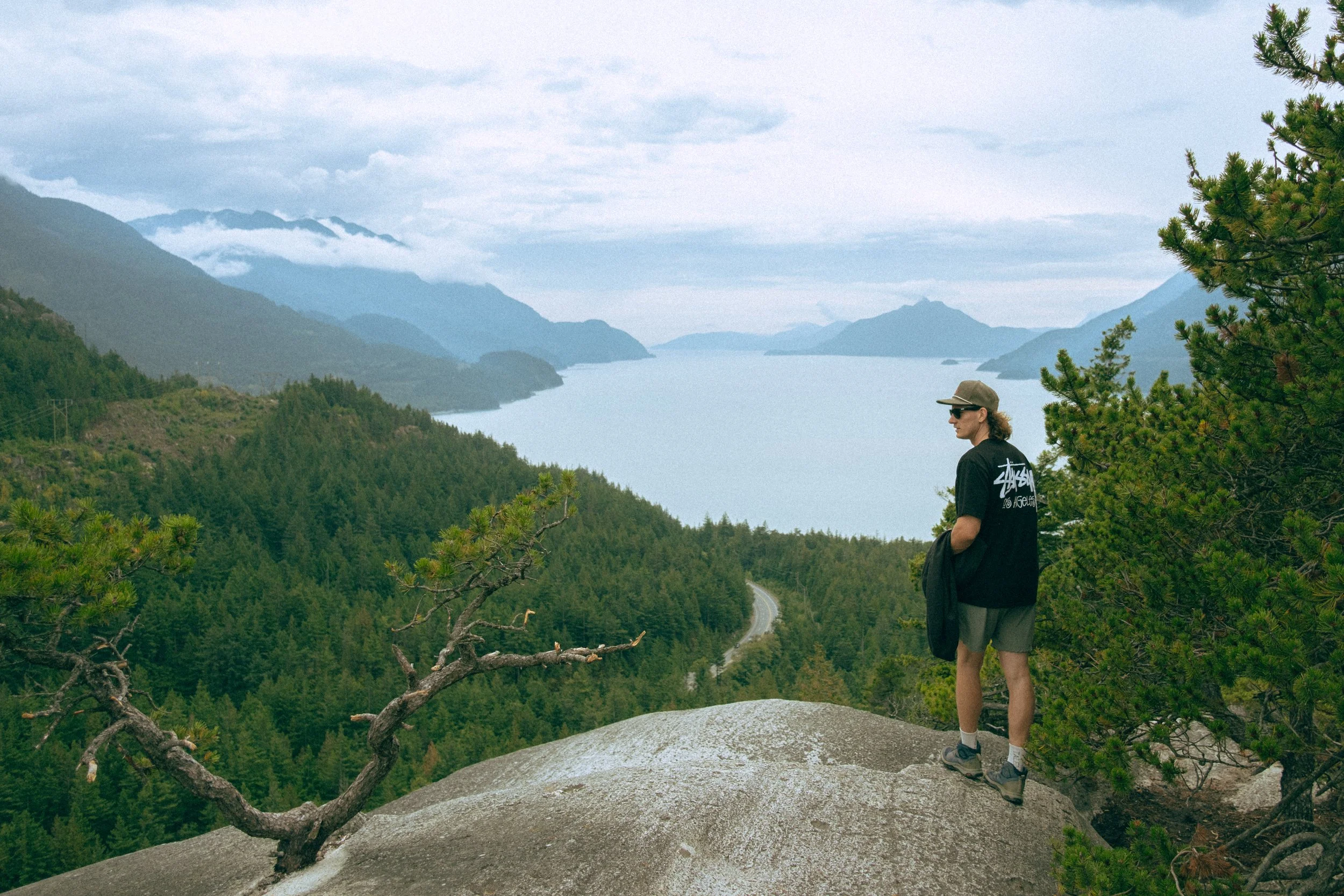 A person standing on a rock overlooking a scenic mountain landscape with a river and forest in the background, wearing sunglasses, a baseball cap, a black shirt, and shorts.