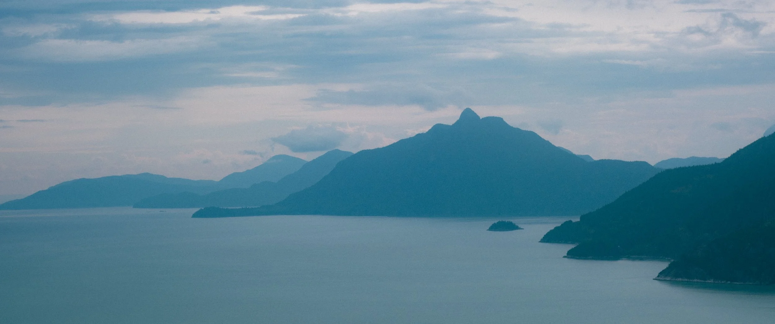 A landscape featuring a large body of water in the foreground, with multiple mountain peaks in the background under a cloudy sky.