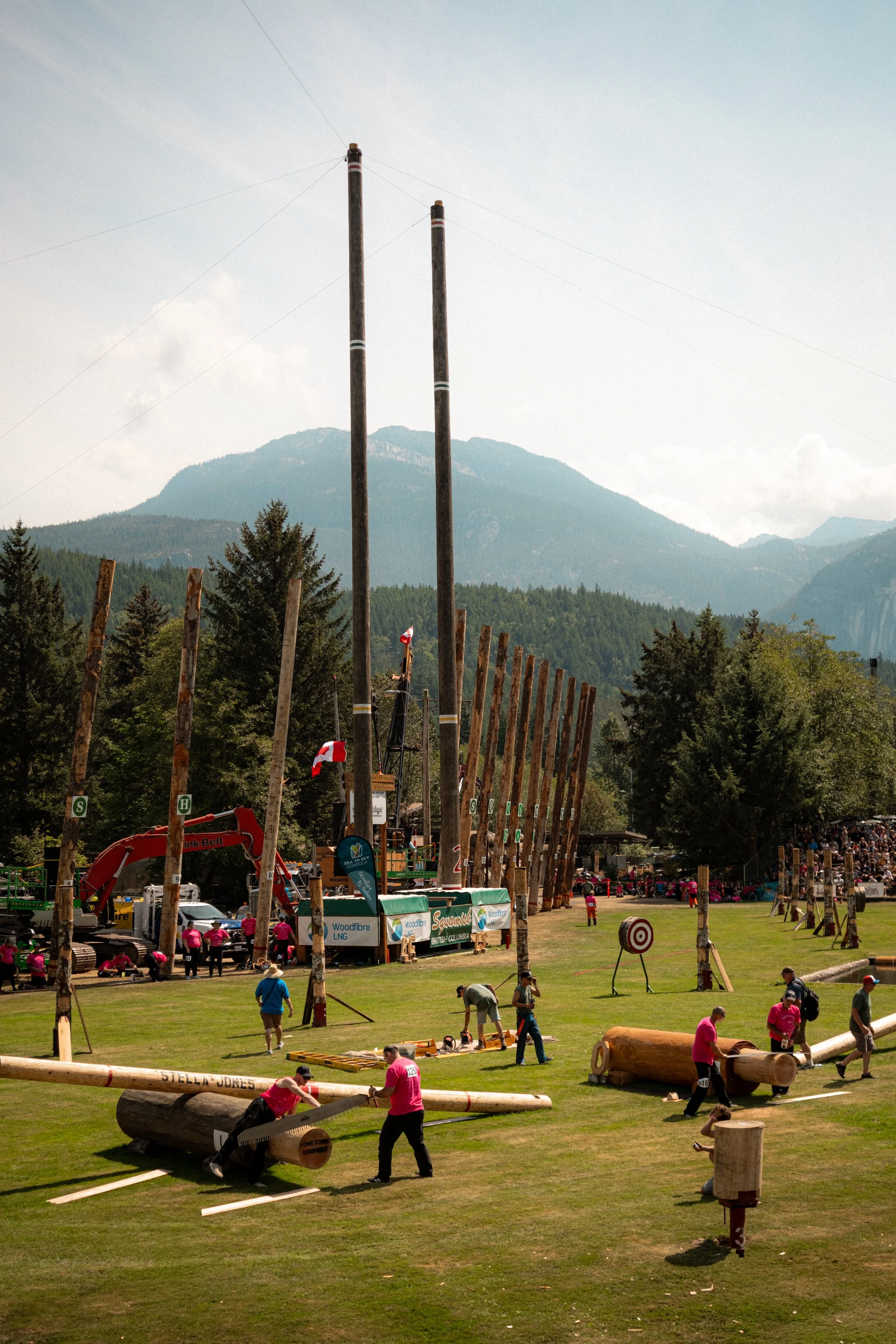 People working on large wooden log structures and poles at an outdoor event with mountains in the background.
