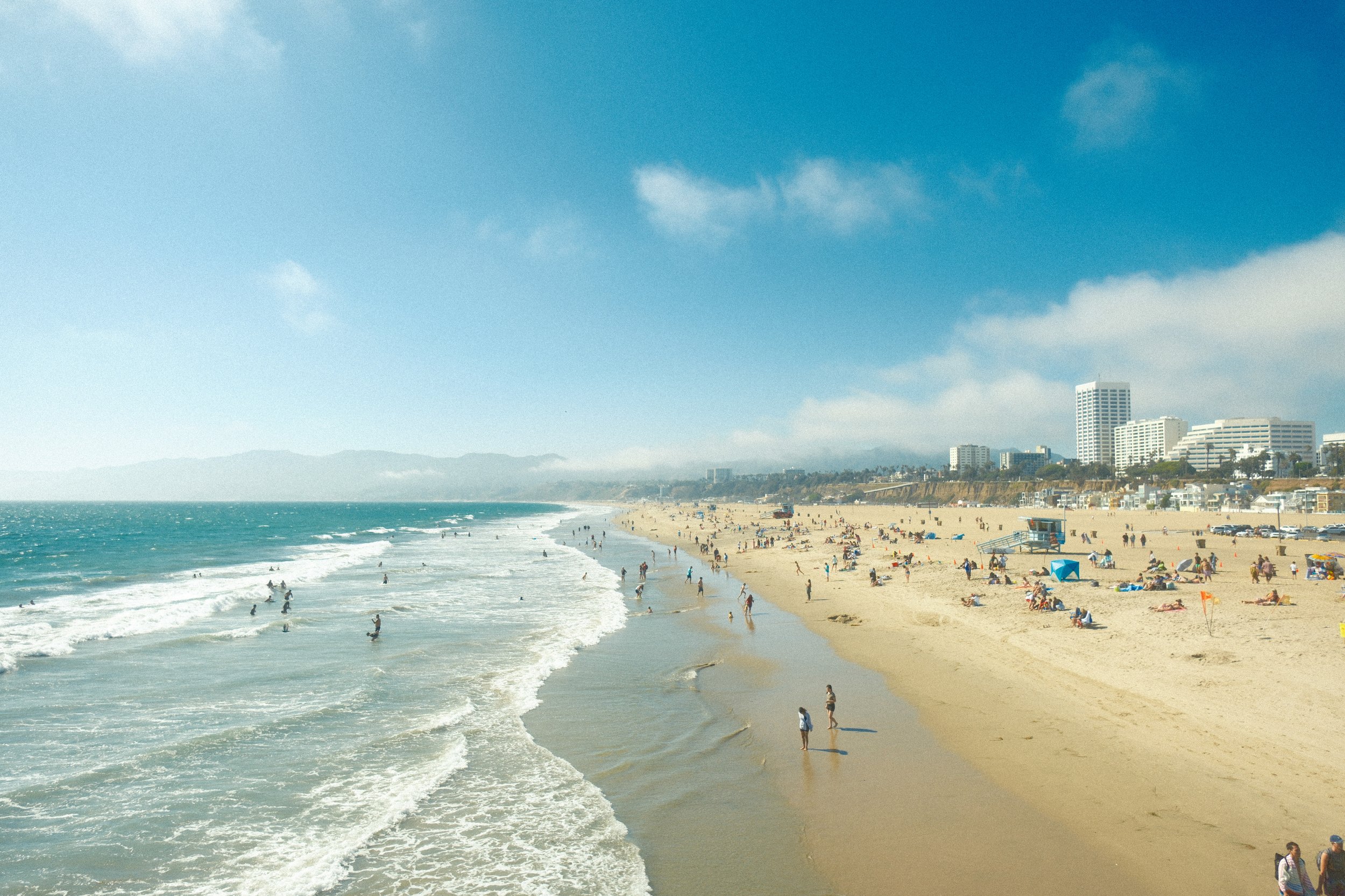 A crowded beach with people relaxing, swimming, and walking along the sandy shore. The ocean waves are crashing onto the beach. In the background, there are several high-rise buildings and a clear blue sky with some clouds.