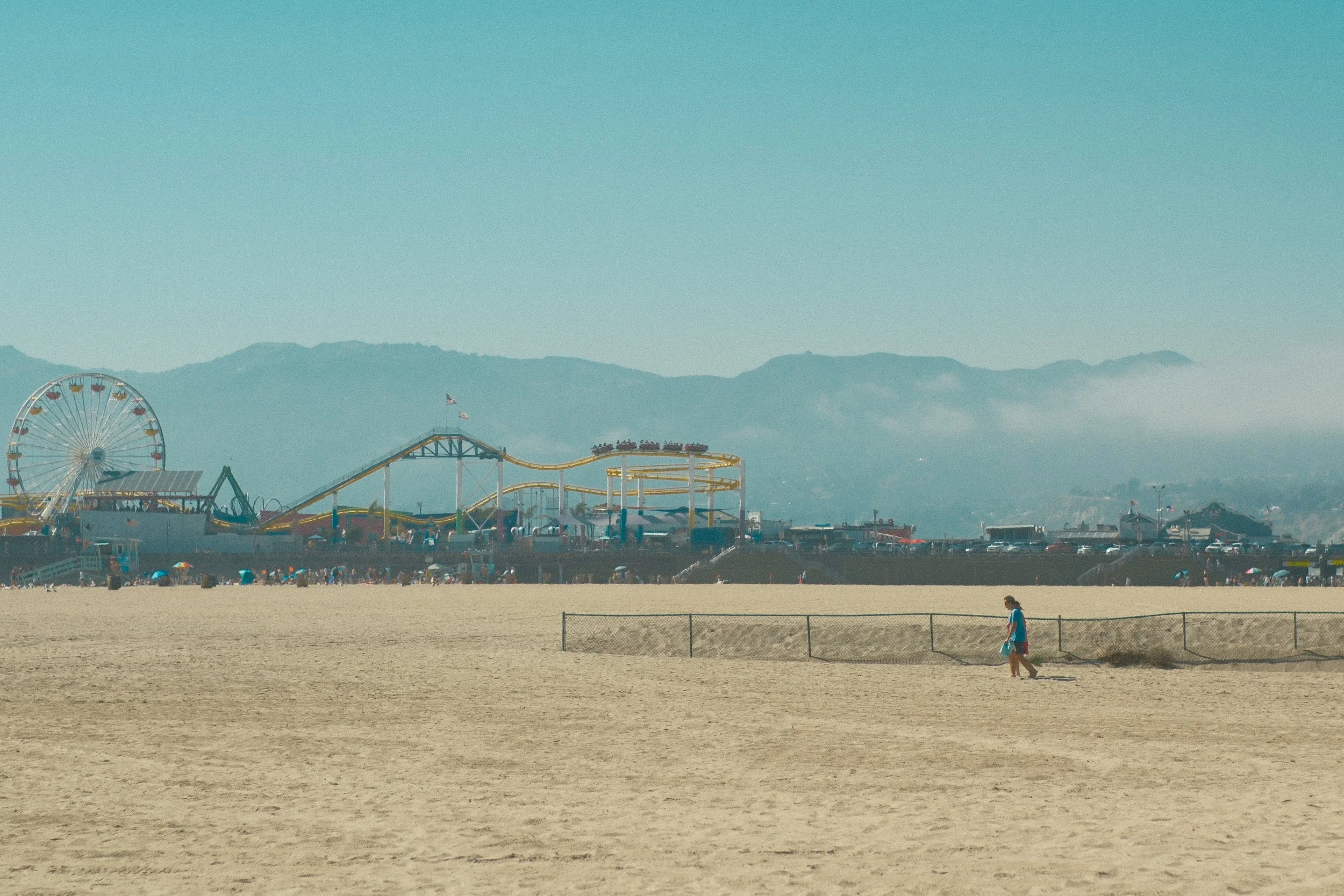 A beach scene with a sandy shore, a woman walking, and an amusement park in the background with a Ferris wheel and roller coaster, mountains, and a clear sky.