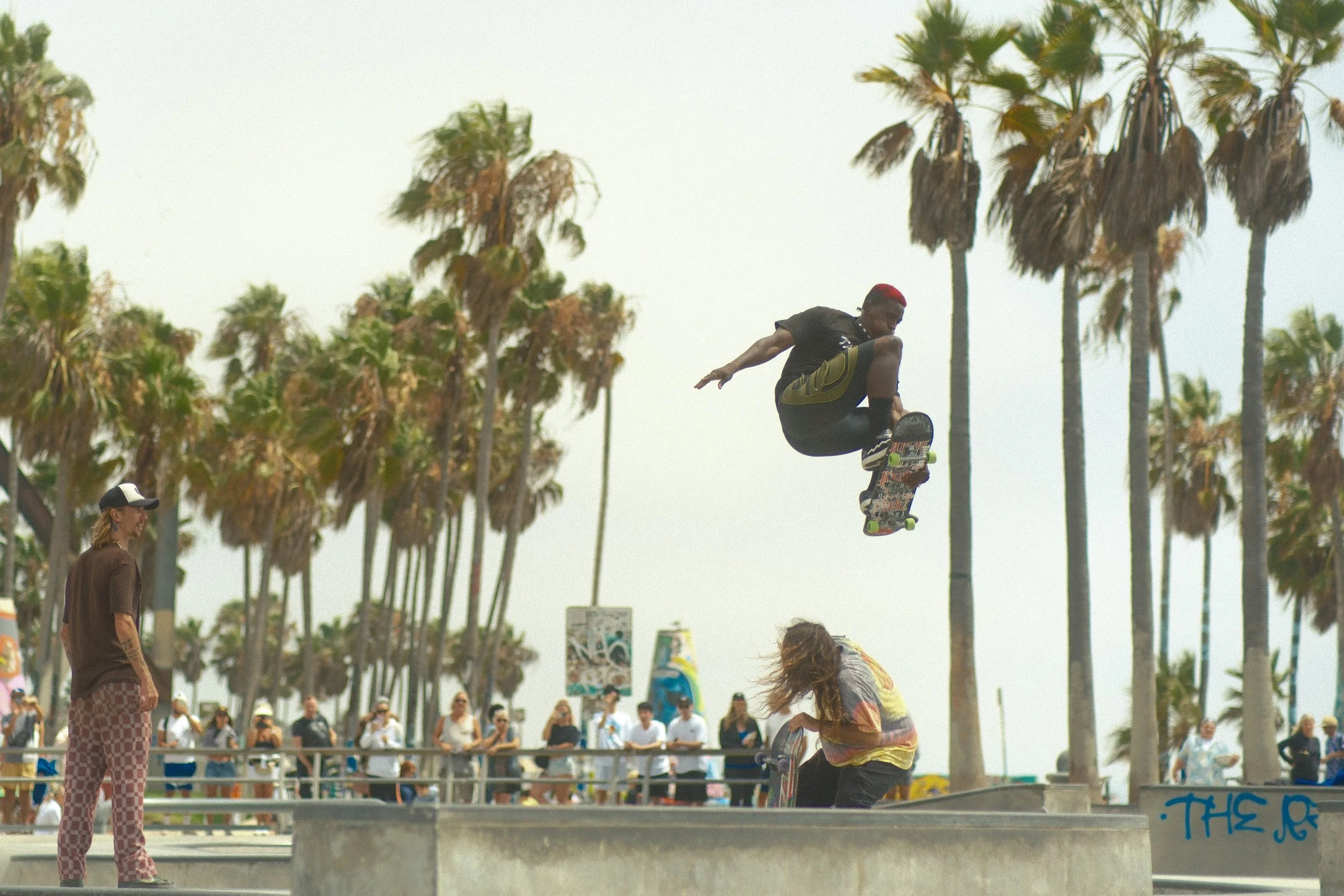 Skateboarder performing a trick in a skatepark with palm trees and spectators in the background.