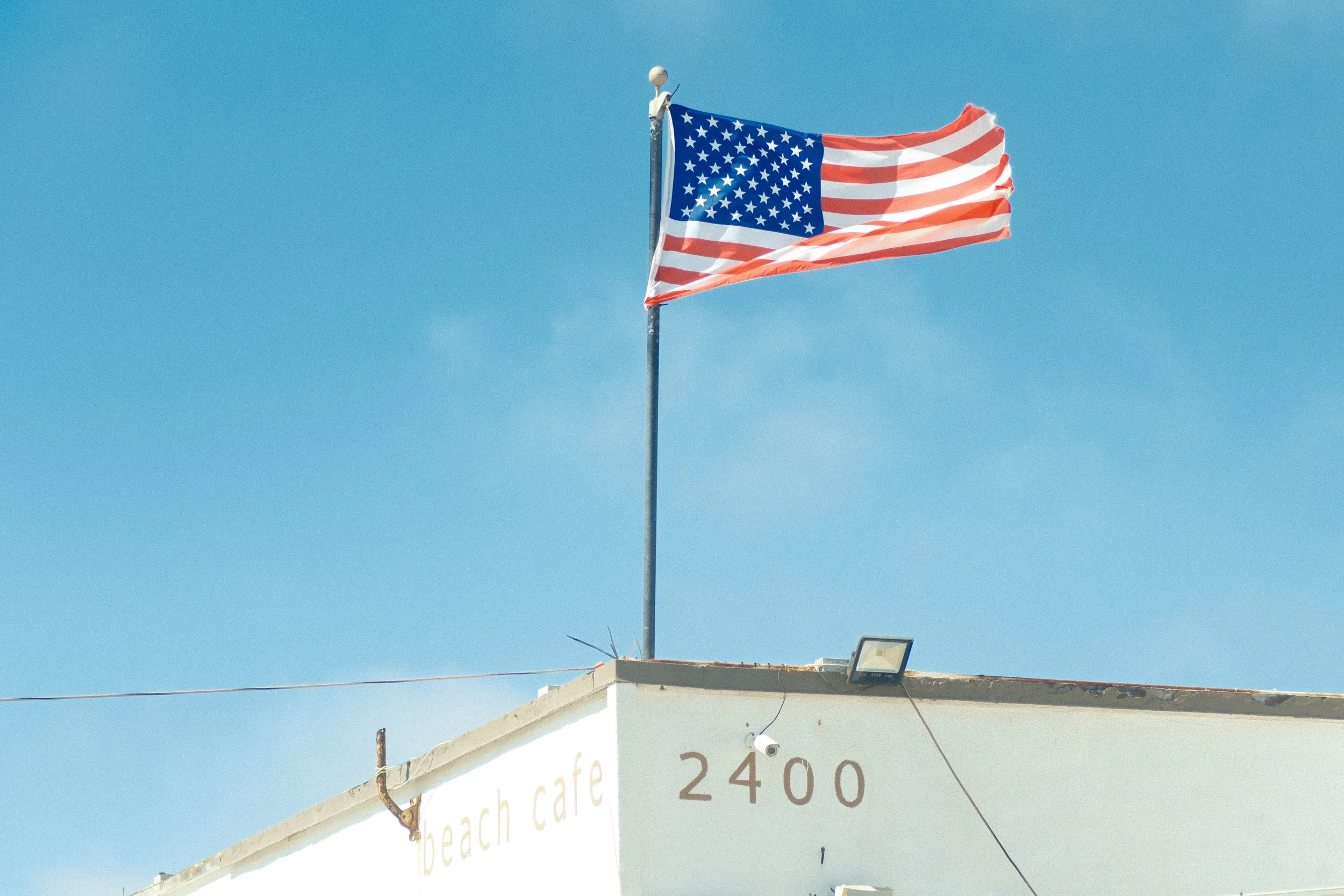 American flag flying on a flagpole above a building with a sign that reads 'beach café 2400' against a clear blue sky.