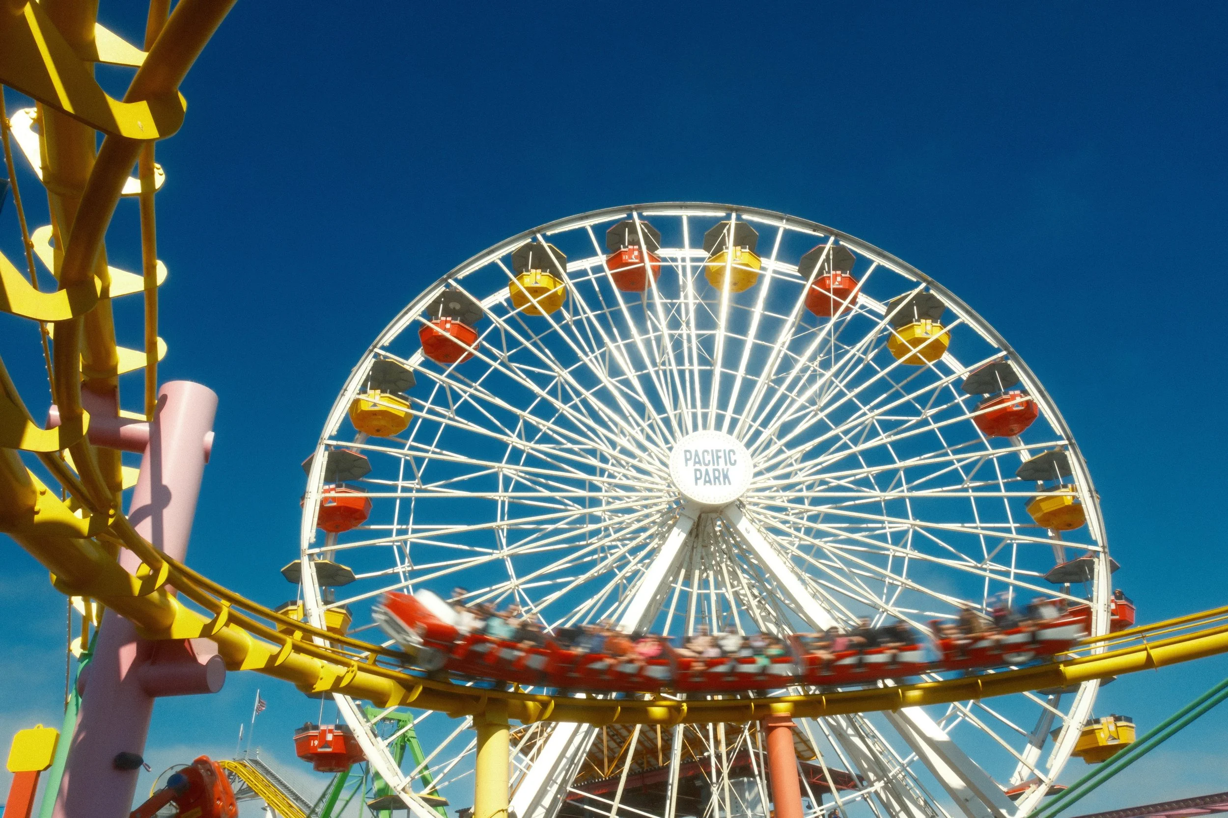 Colorful Ferris wheel and roller coaster at an amusement park against a clear blue sky.