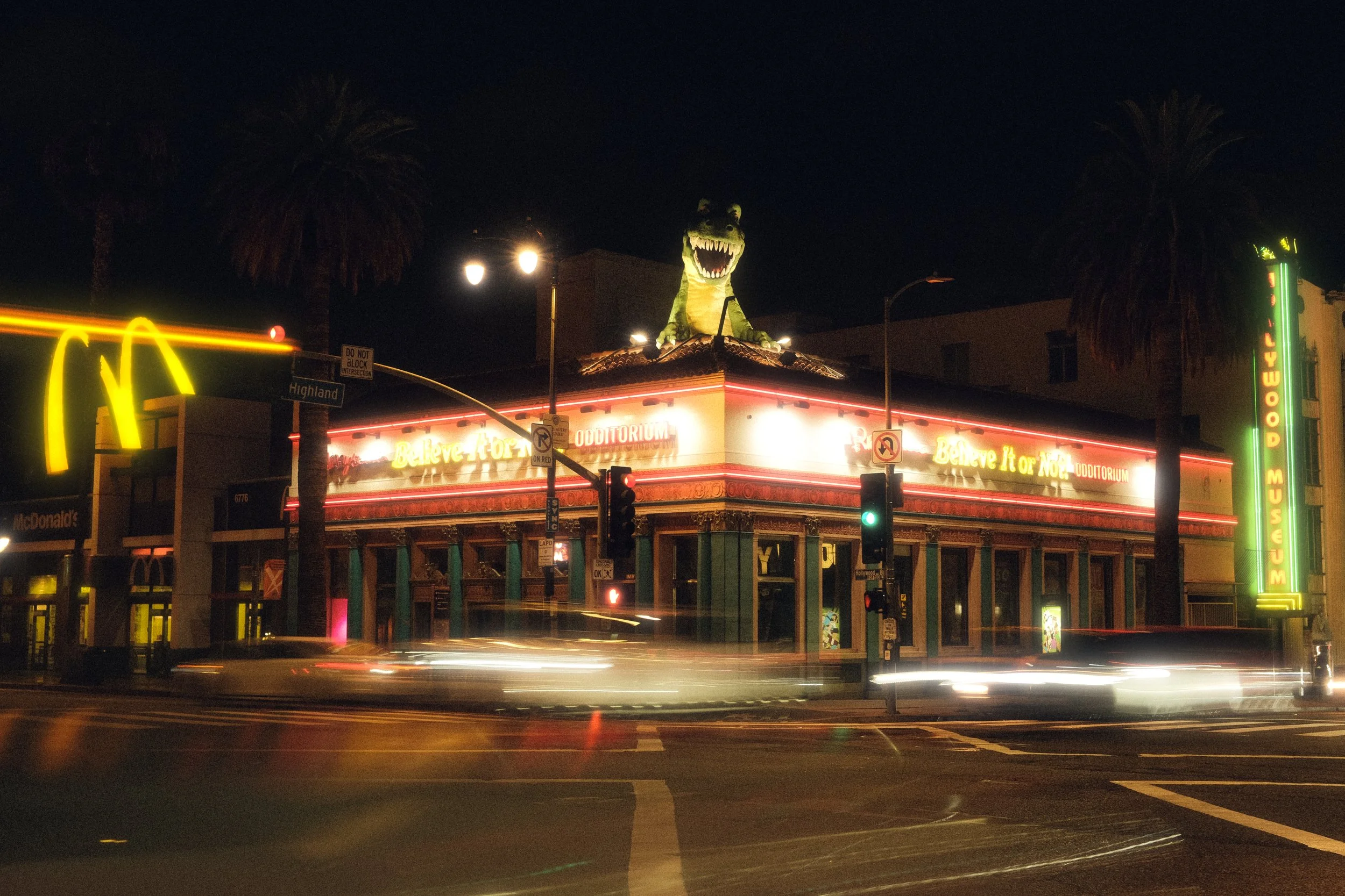 Nighttime scene of a downtown street corner featuring a well-lit building with a dinosaur sculpture on its roof, illuminated signs, palm trees, and streaks of car lights from long exposure.
