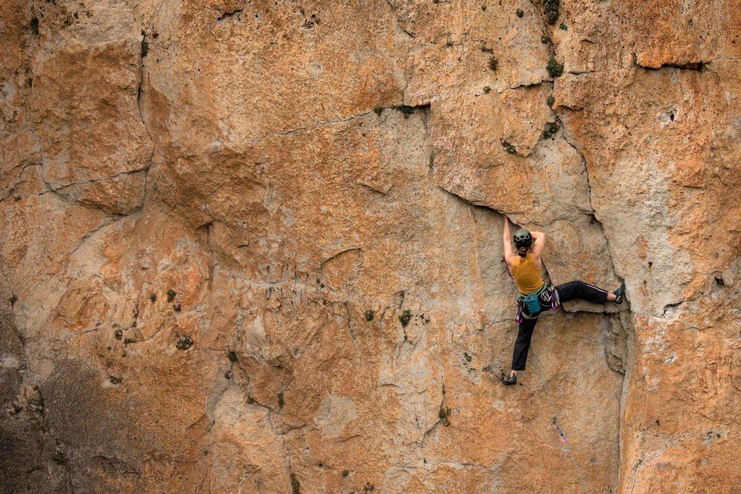 A person rock climbing on a large, textured, orange-brown stone wall. The climber is wearing a helmet, a yellow shirt, black pants, and climbing gear, and is secured with a harness and rope.