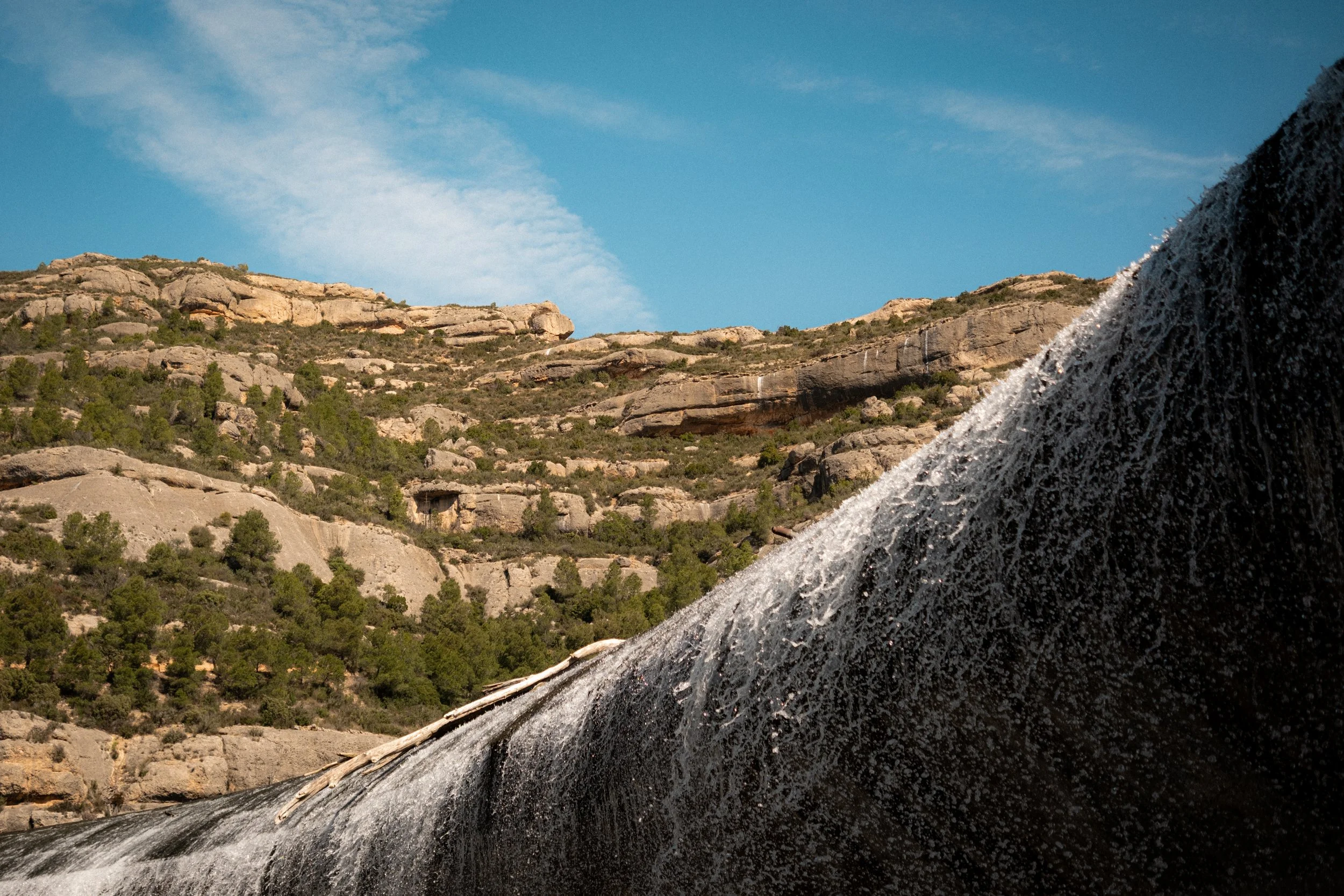 A rocky desert landscape with a waterfall flowing down the rocks, under a blue sky with some clouds.