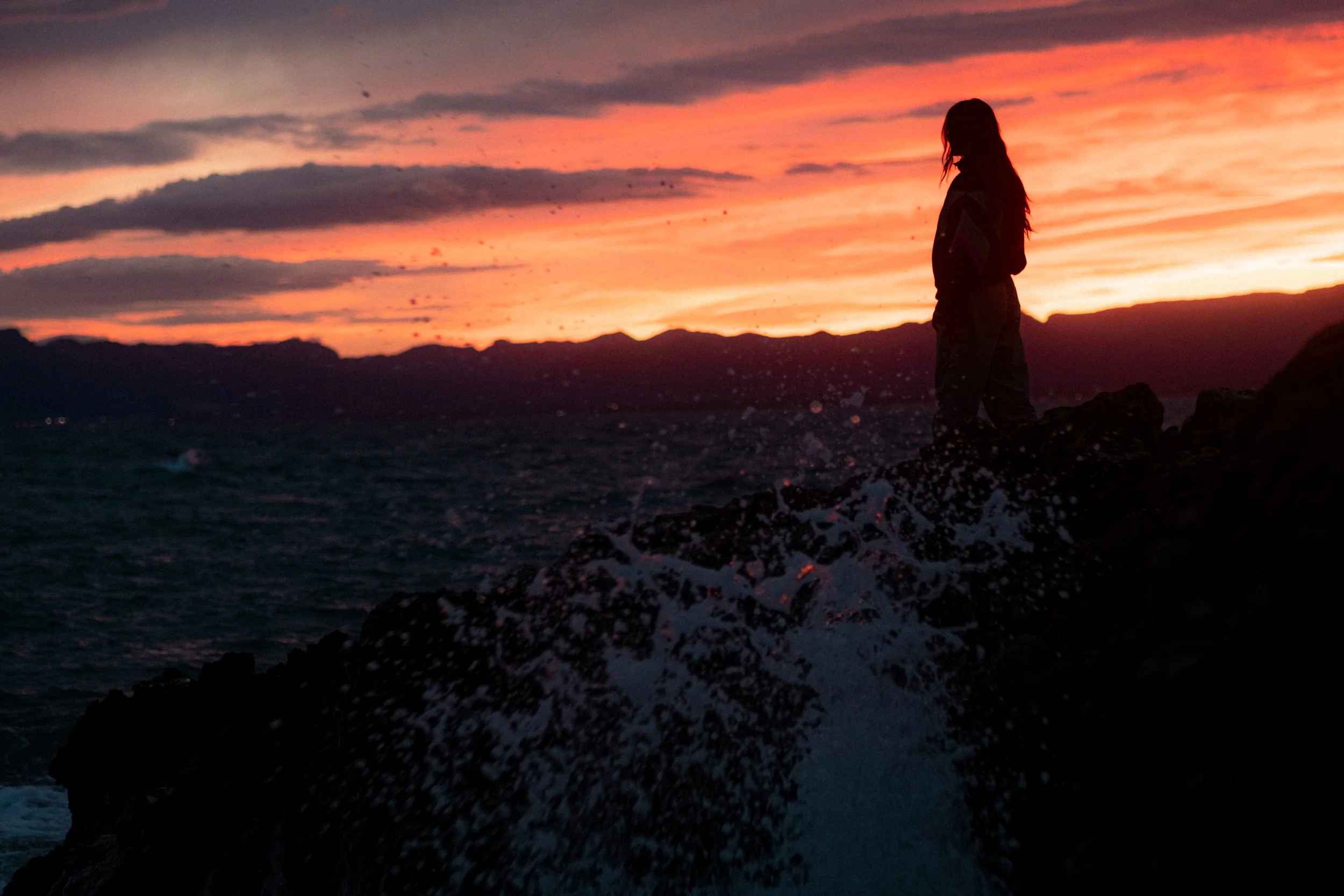 A person standing on rocks near the water during a sunset with colorful sky and clouds.