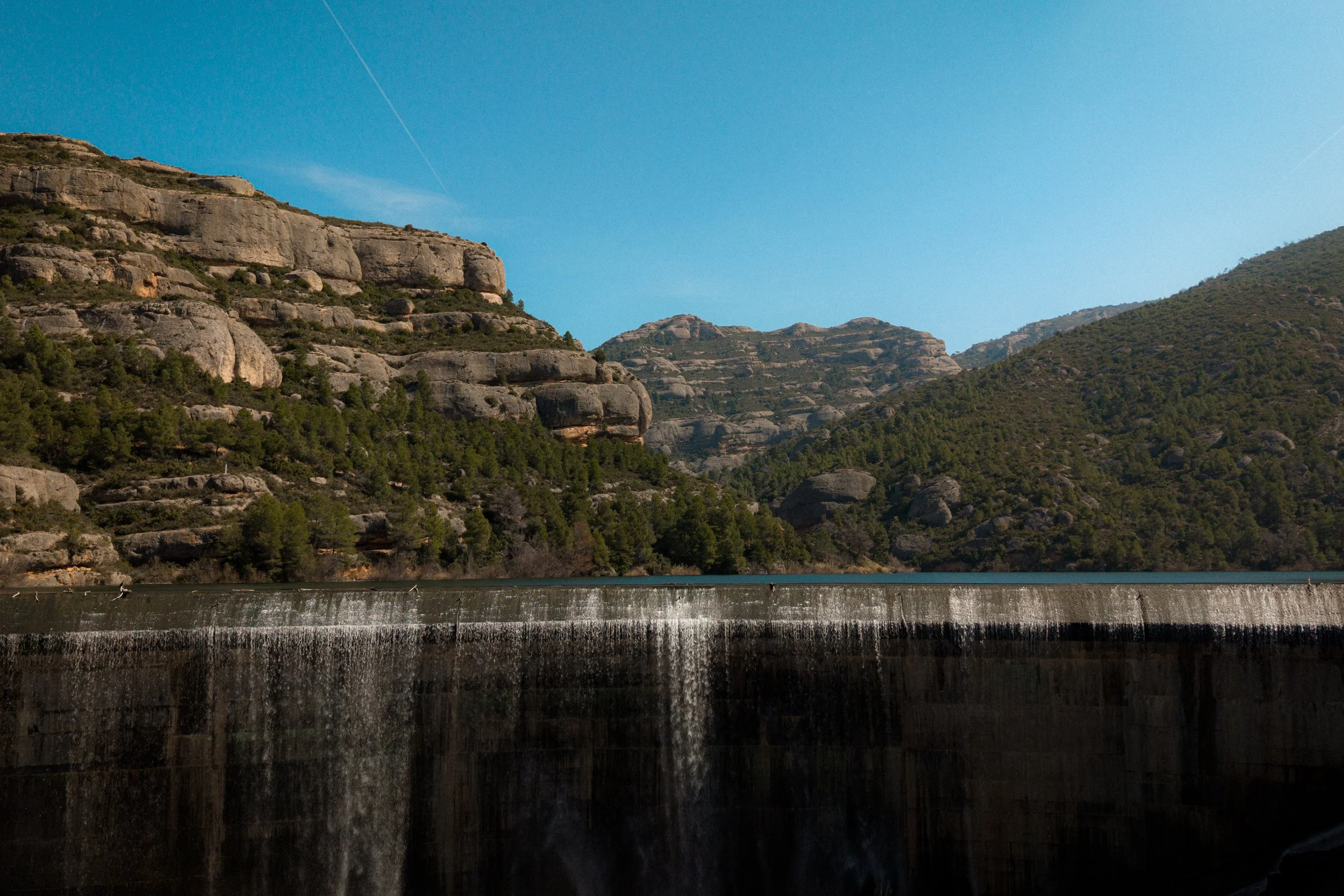 A scenic view of mountains with rocky cliffs and green trees beside a body of water, under a clear blue sky with contrails.