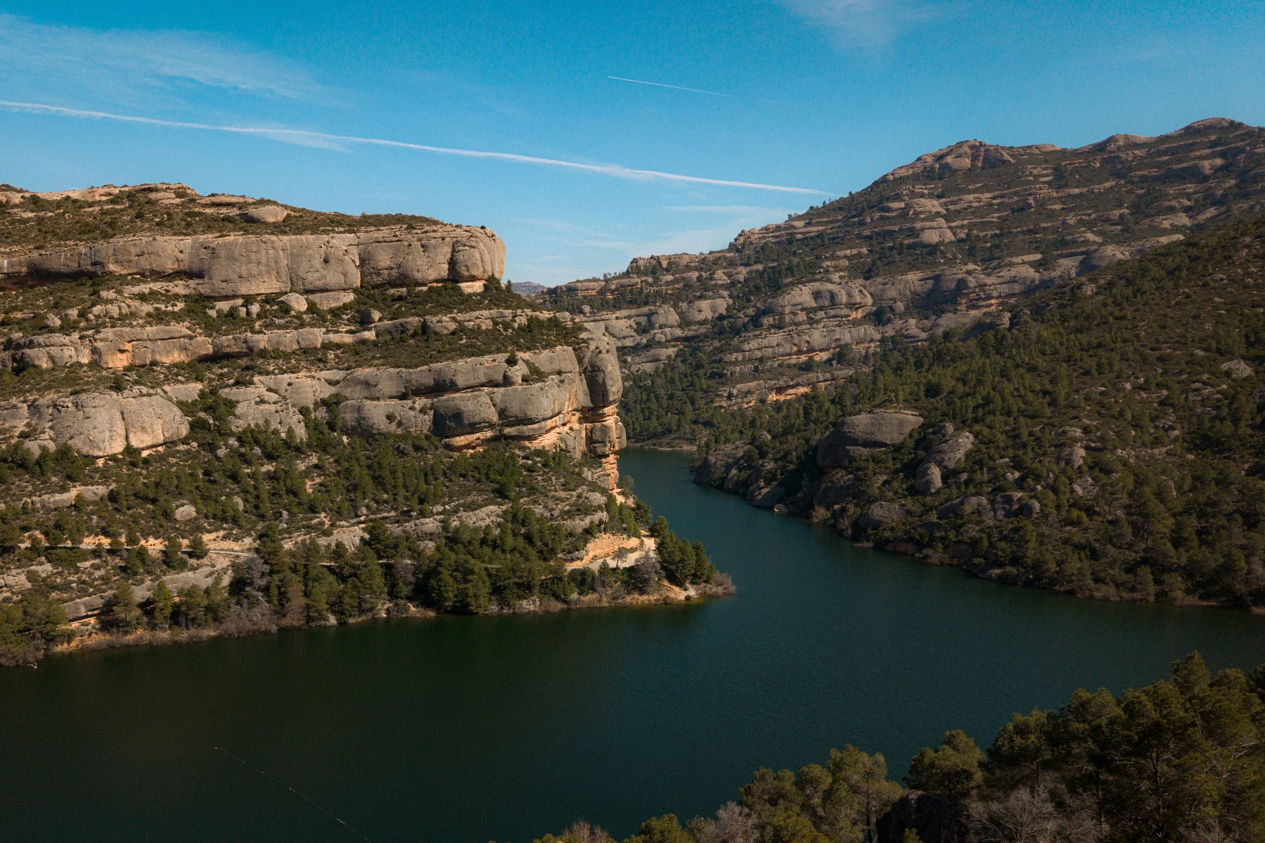 A scenic view of a river flowing through canyon with rocky cliffs and green trees under a blue sky with a few clouds and a contrail.