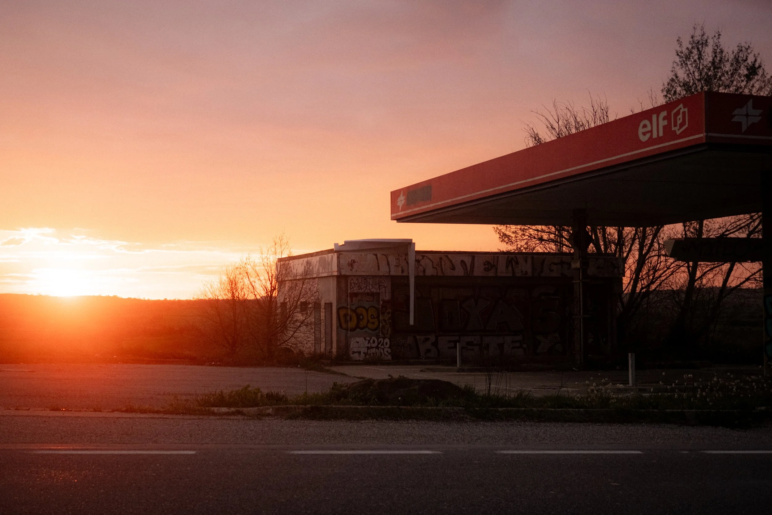 A sunset scene showing a gas station with an orange and red sign labeled 'elf' next to a graffiti-covered building and leafless trees in the background.