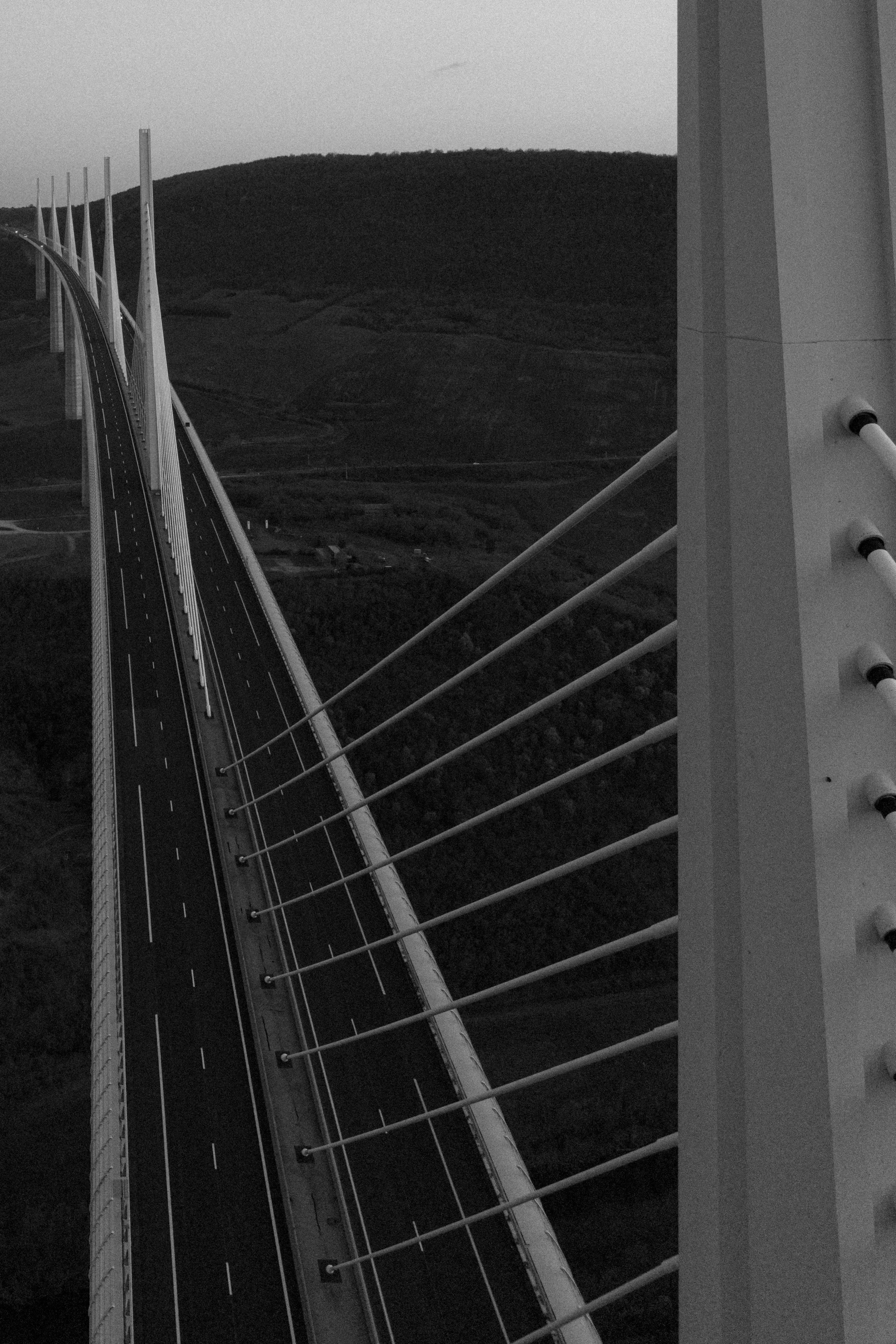 Black and white photo of a bridge with tall, curved support towers and suspension cables over a hilly landscape.