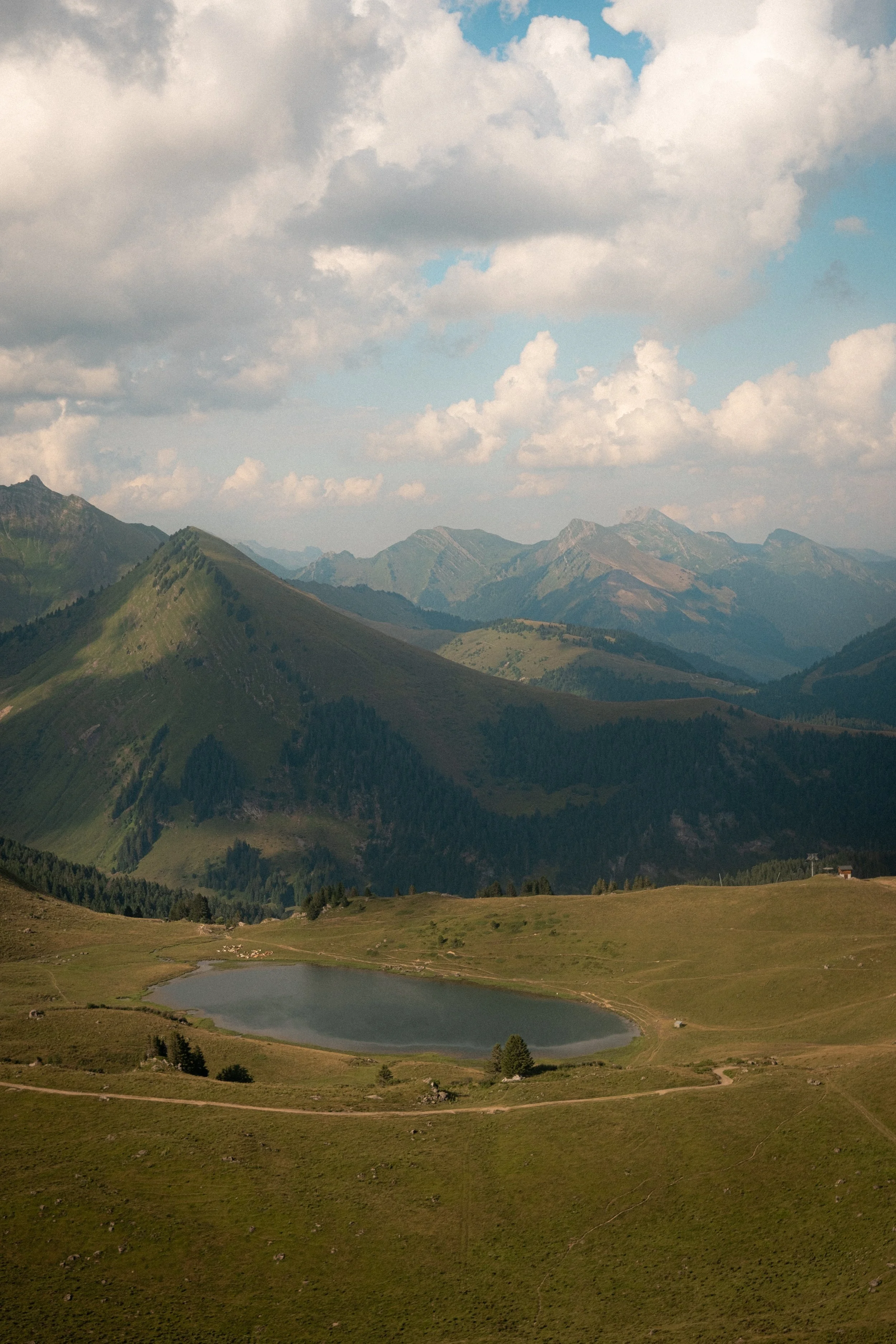 Scenic view of a mountain range with a small lake in the foreground, green hills, and a partly cloudy sky.